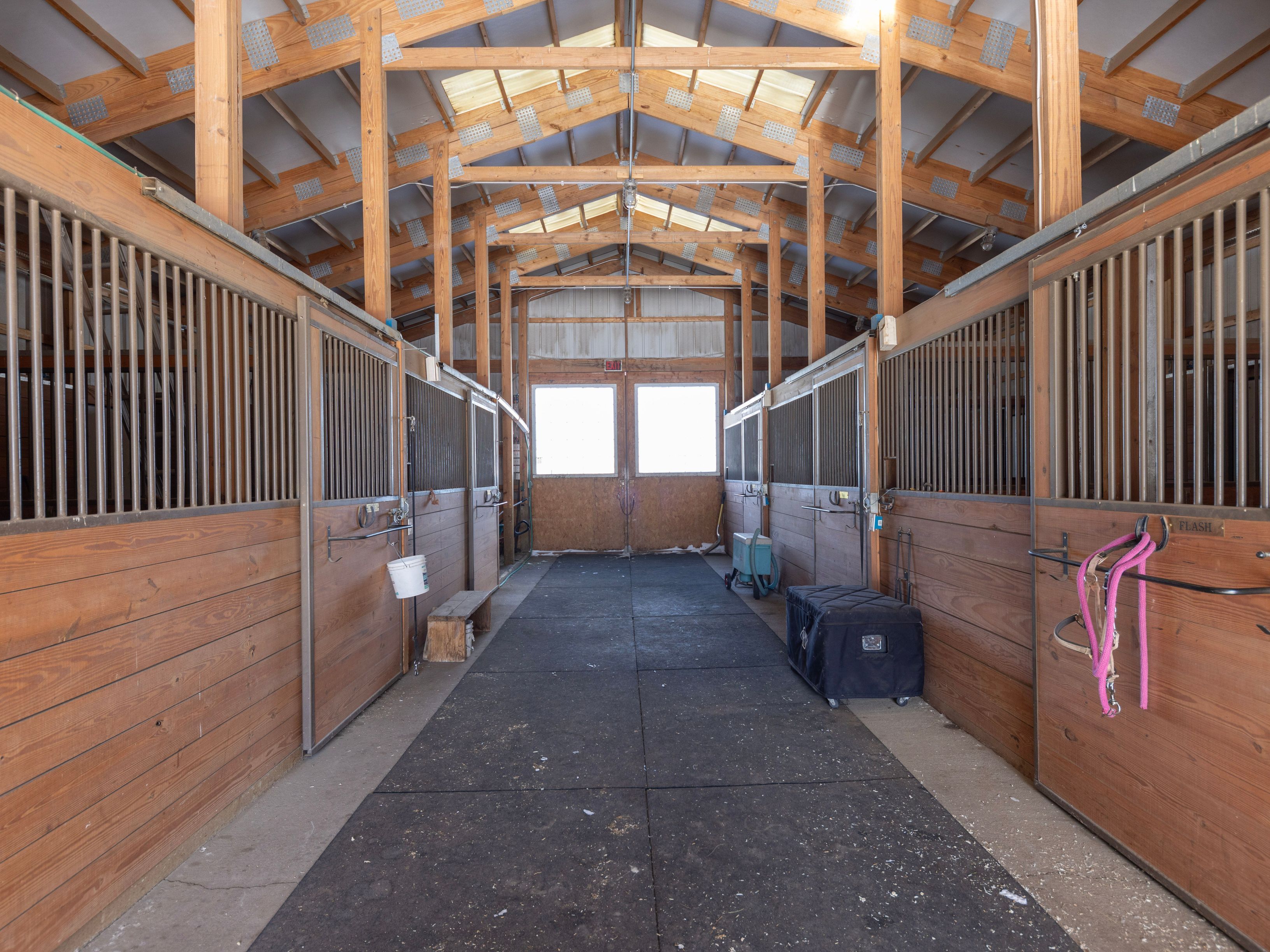 This image captures the interior aisle of a well-maintained horse stable, featuring rows of wooden stalls with metal bars on either side. The perspective is a centered, eye-level shot looking down the rubber-matted walkway toward a pair of bright, light-filled double doors at the far end. The exposed wooden rafters and trusses overhead add a rustic, structural charm to the functional equestrian space.