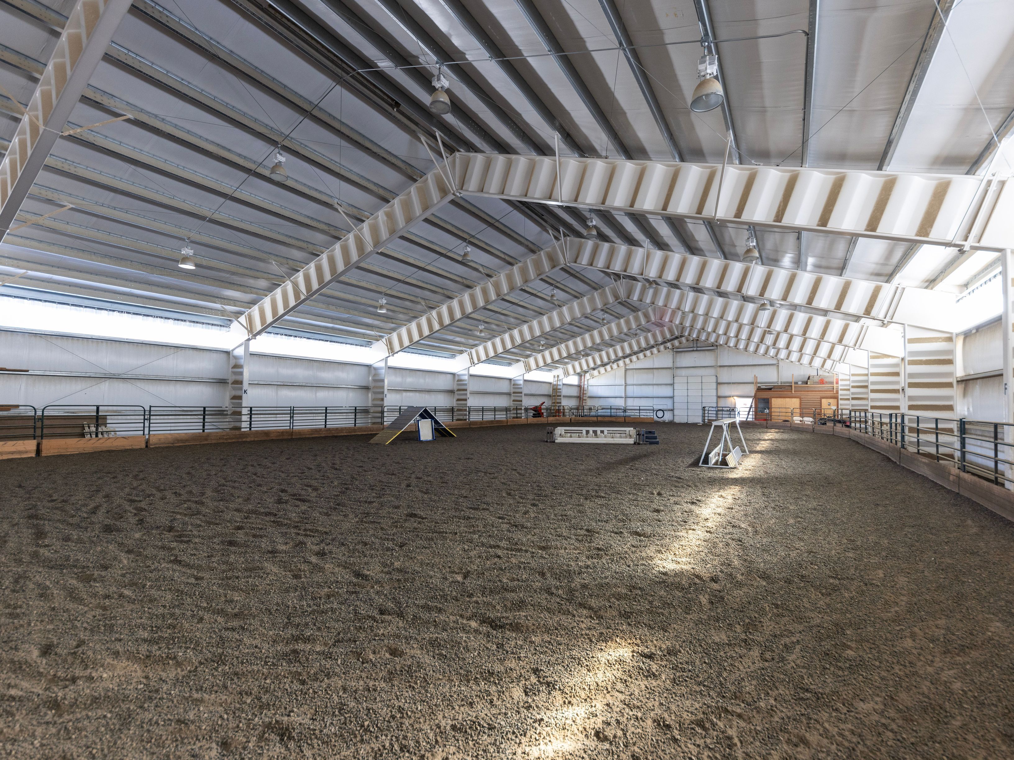 This image showcases a large, indoor equestrian riding arena with a dirt floor and high, vaulted ceilings supported by exposed metal trusses. The space is equipped with various agility obstacles, suggesting it is used for horse or dog training, and is illuminated by multiple overhead industrial-style lights. The perspective is wide-angle, capturing the expansive, functional nature of the facility.
