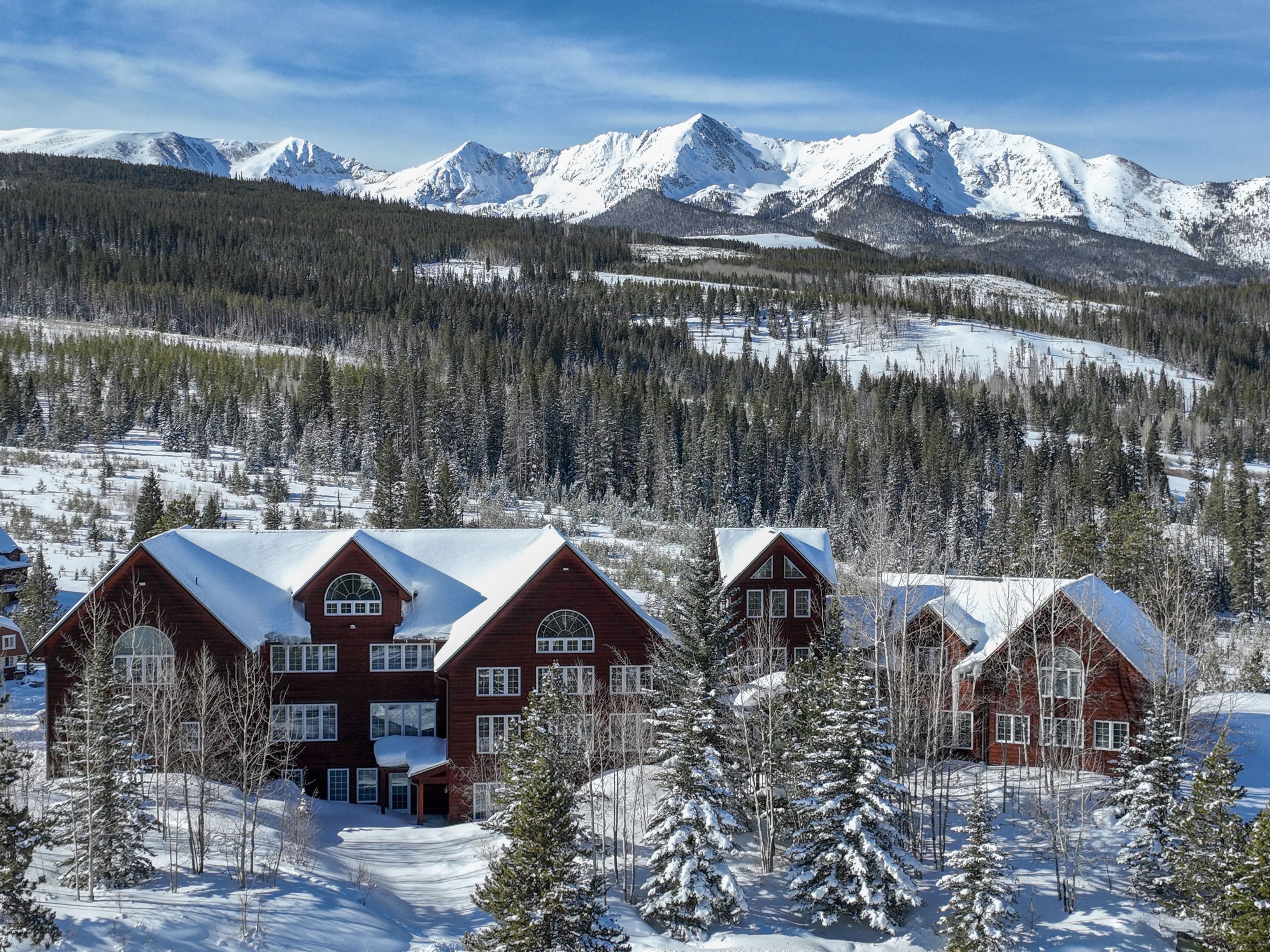 This stunning aerial shot captures a large, multi-story mountain lodge nestled within a dense, snow-covered pine forest. The home features classic rustic architecture with dark wood siding, numerous windows, and snow-dusted gabled roofs, set against a breathtaking backdrop of jagged, snow-capped mountain peaks under a clear blue sky. The perspective emphasizes the property's secluded, serene location and its harmonious integration with the rugged alpine landscape.