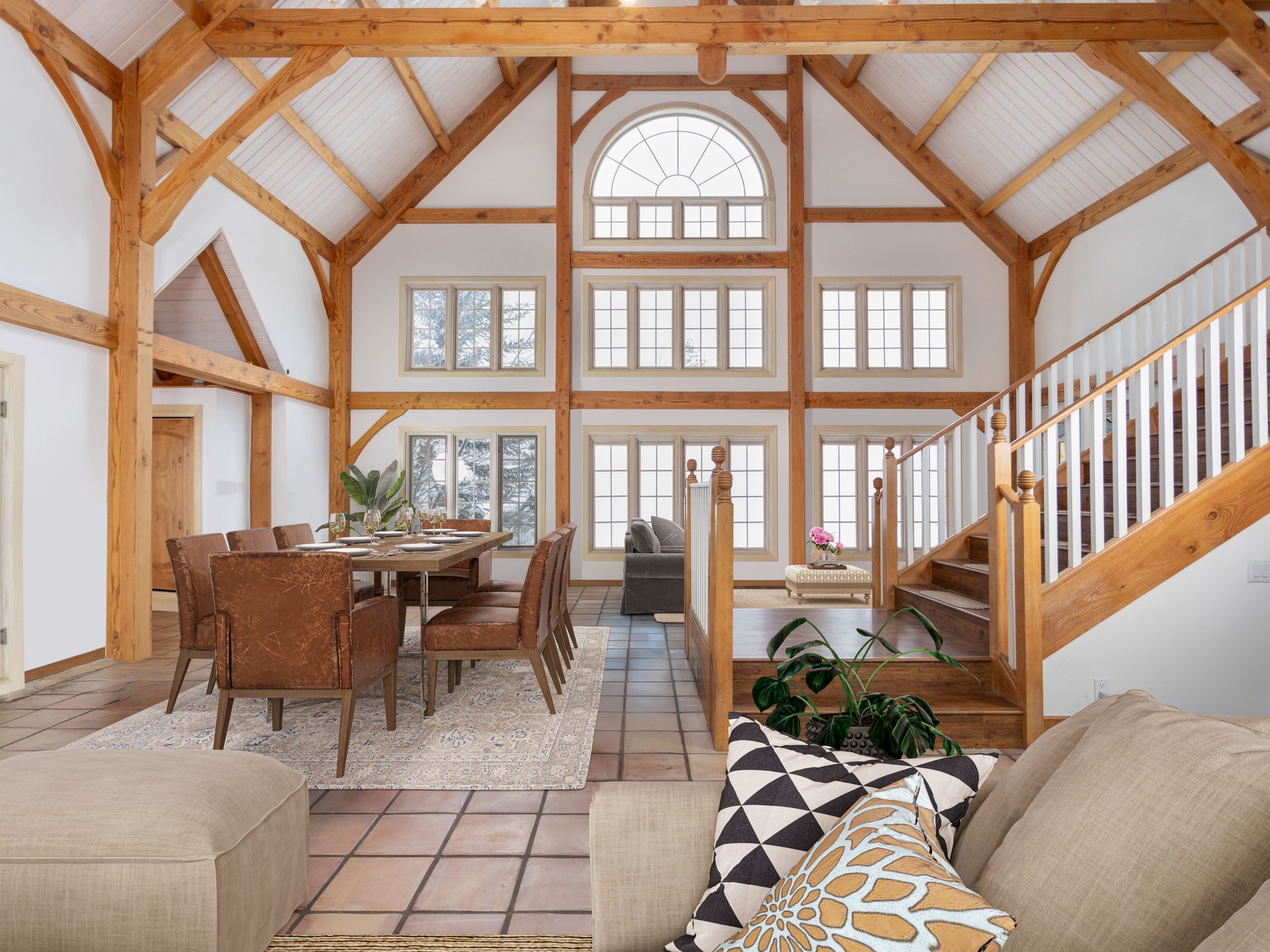This expansive, open-concept living and dining area features a stunning timber-frame construction with vaulted ceilings and exposed wooden beams. The space is flooded with natural light from a large, multi-tiered window wall, highlighting the rustic elegance of the wood architecture and the warm, earthy tones of the tile flooring. A formal dining set sits on a patterned rug, while a staircase with white railings leads to the upper level, creating a grand and inviting atmosphere.