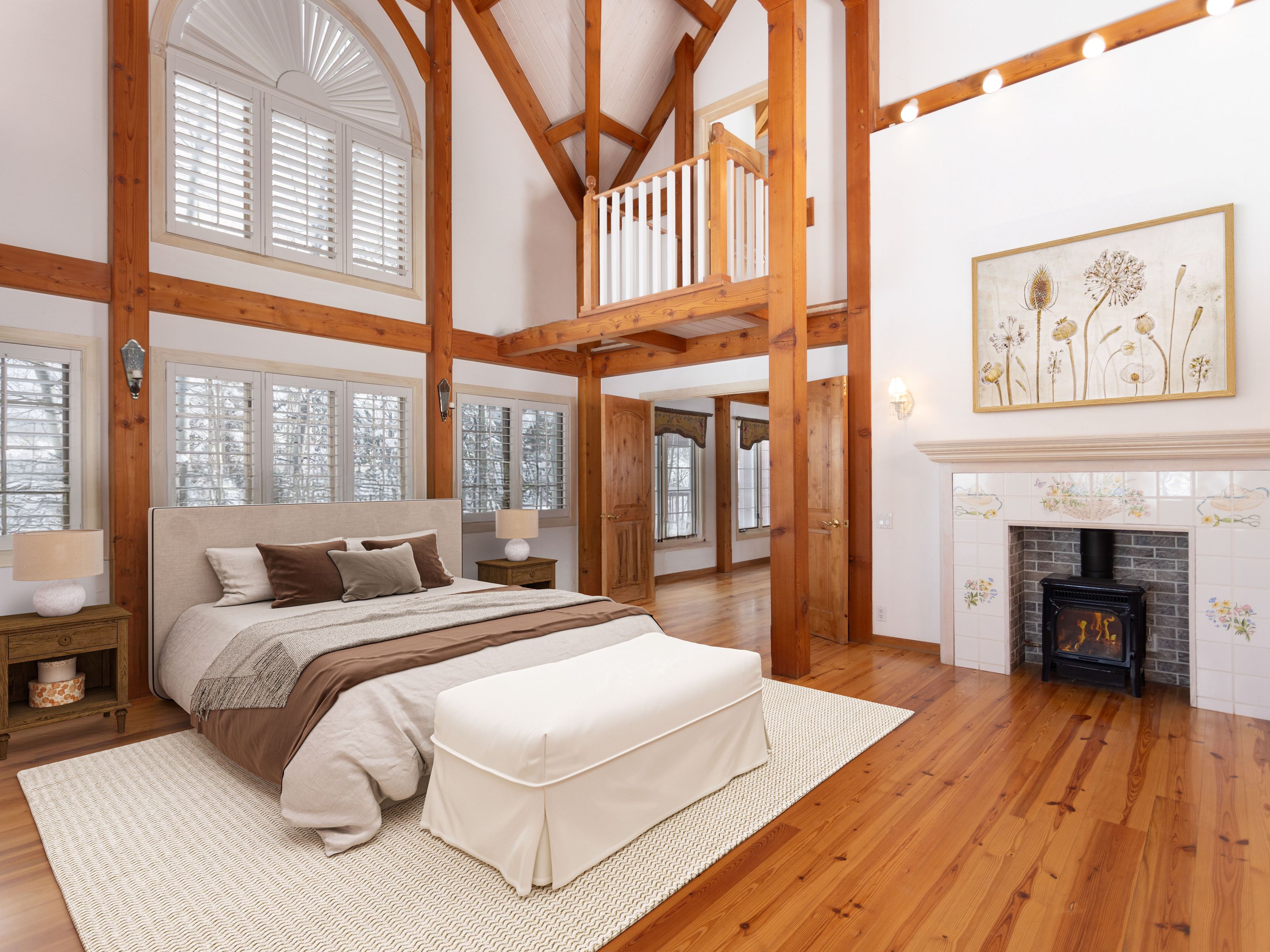 This spacious primary bedroom features a dramatic vaulted ceiling with exposed timber beams and a lofted balcony overlooking the room. The space is anchored by a cozy fireplace with a wood-burning stove and a large, neutral-toned bed, all set against warm hardwood floors and expansive windows with plantation shutters. The overall aesthetic is a sophisticated blend of rustic timber-frame architecture and elegant, comfortable interior design.