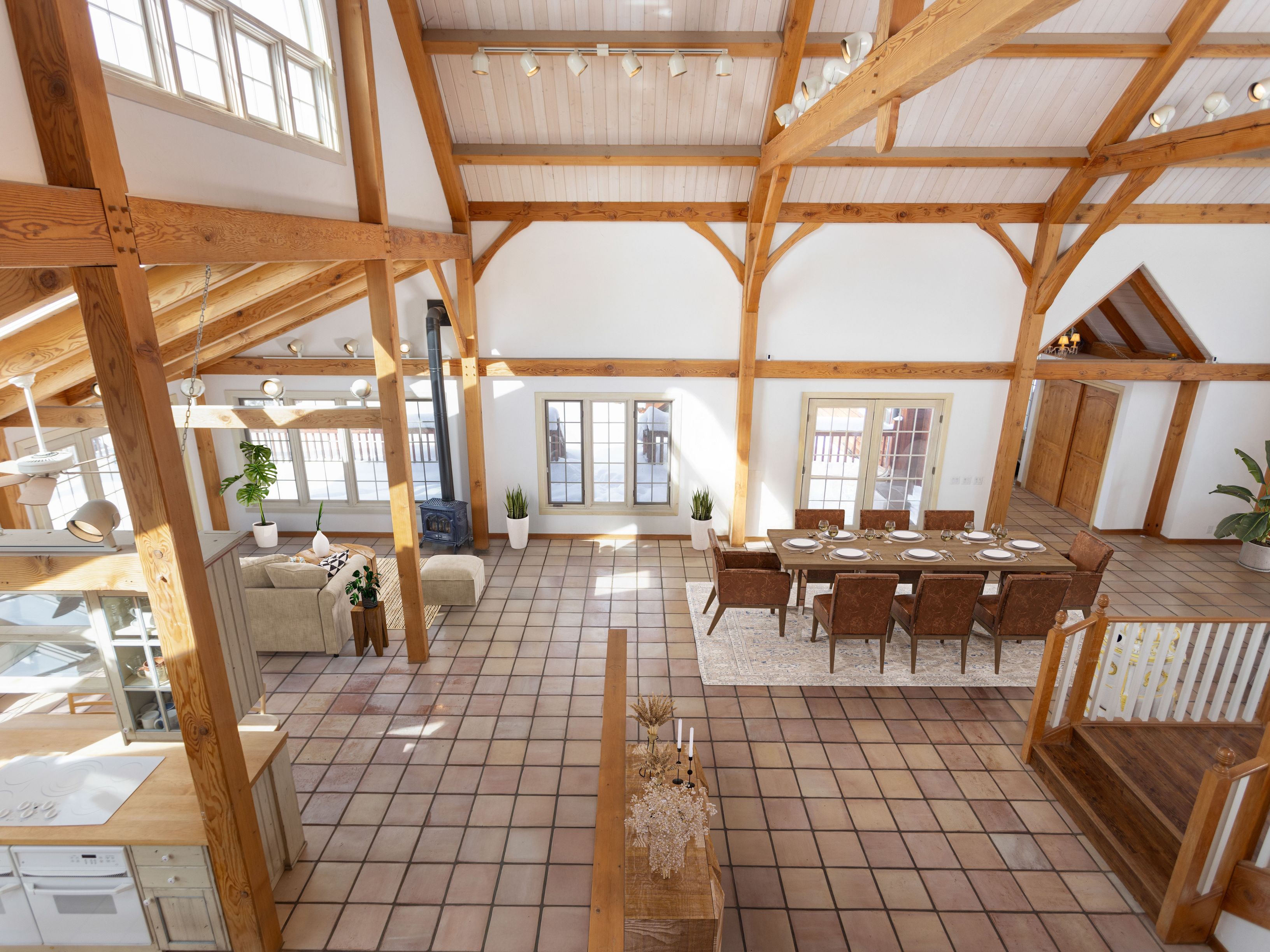 This high-angle, wide-shot perspective captures a spacious, open-concept living and dining area defined by dramatic timber-frame vaulted ceilings and rustic exposed beams. The room features a neutral color palette with terracotta-style tiled flooring, a cozy seating area with a wood-burning stove, and a formal dining set, all bathed in natural light from large windows and glass doors. The architectural design creates an airy, lodge-like atmosphere that seamlessly blends rustic charm with expansive, modern living space.
