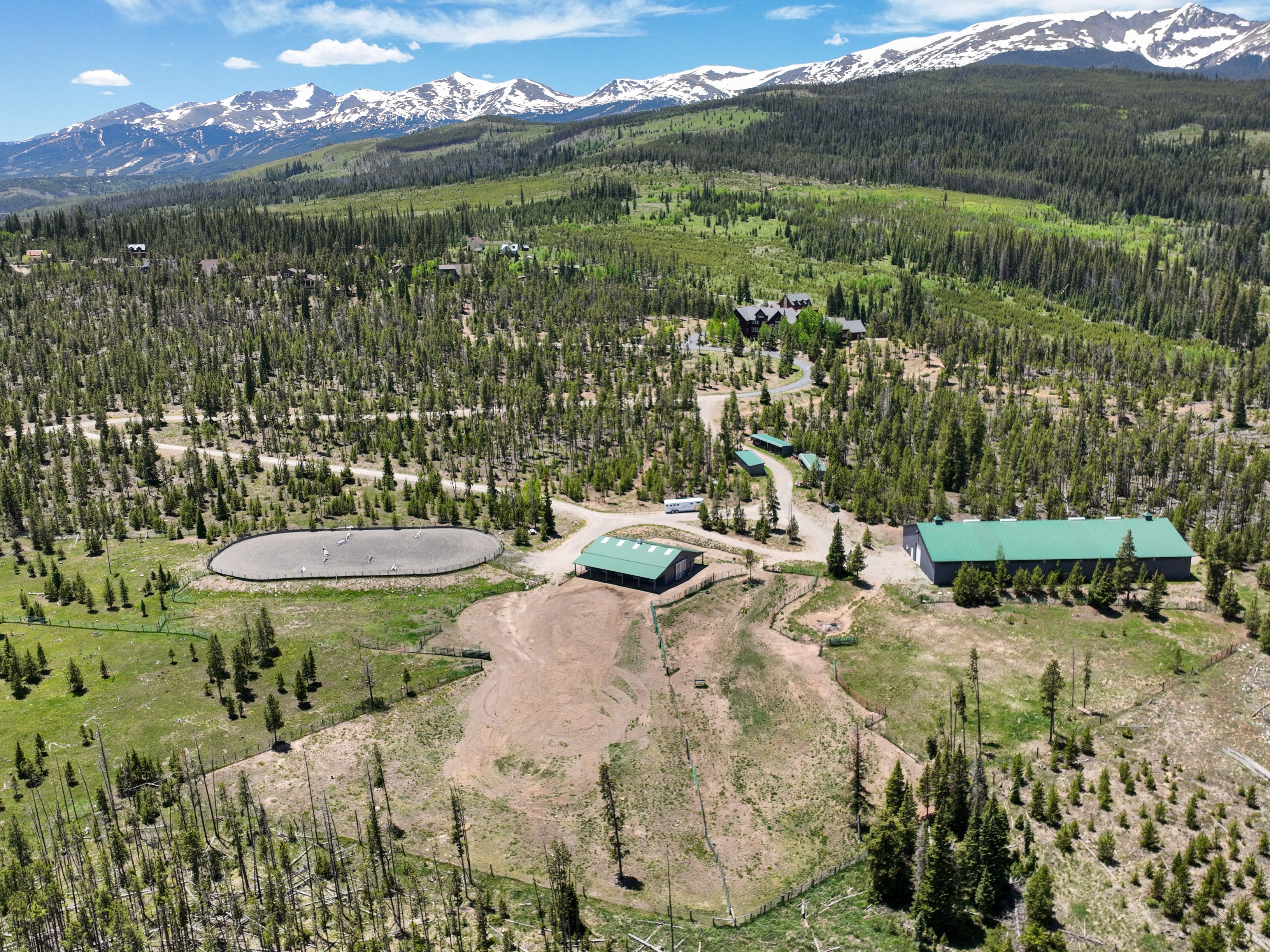 This expansive aerial view captures a sprawling mountain property featuring a large equestrian facility with a riding arena and multiple barns. The estate is nestled within a dense, lush forest, set against a dramatic backdrop of snow-capped mountain peaks under a clear blue sky. The property layout emphasizes a rural, high-altitude lifestyle with significant acreage and private infrastructure.