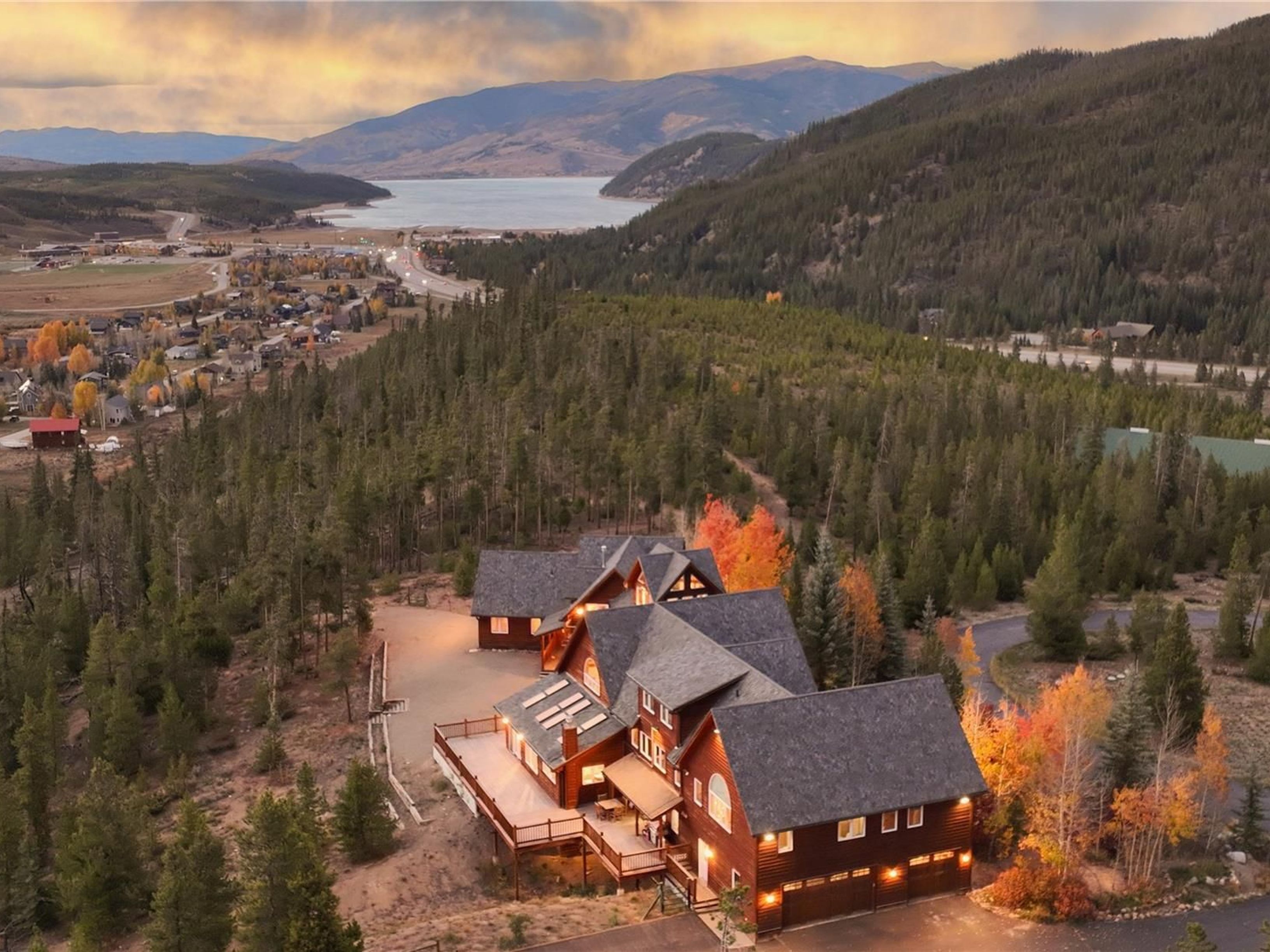 This stunning aerial perspective captures a large, multi-level log-style mountain home nestled within a dense evergreen forest. The property features an expansive wooden deck, a multi-car garage, and a complex roofline, all set against a breathtaking backdrop of a distant lake and rolling mountains at sunset. The warm, golden hour lighting enhances the rustic charm of the architecture, creating an inviting and luxurious atmosphere.