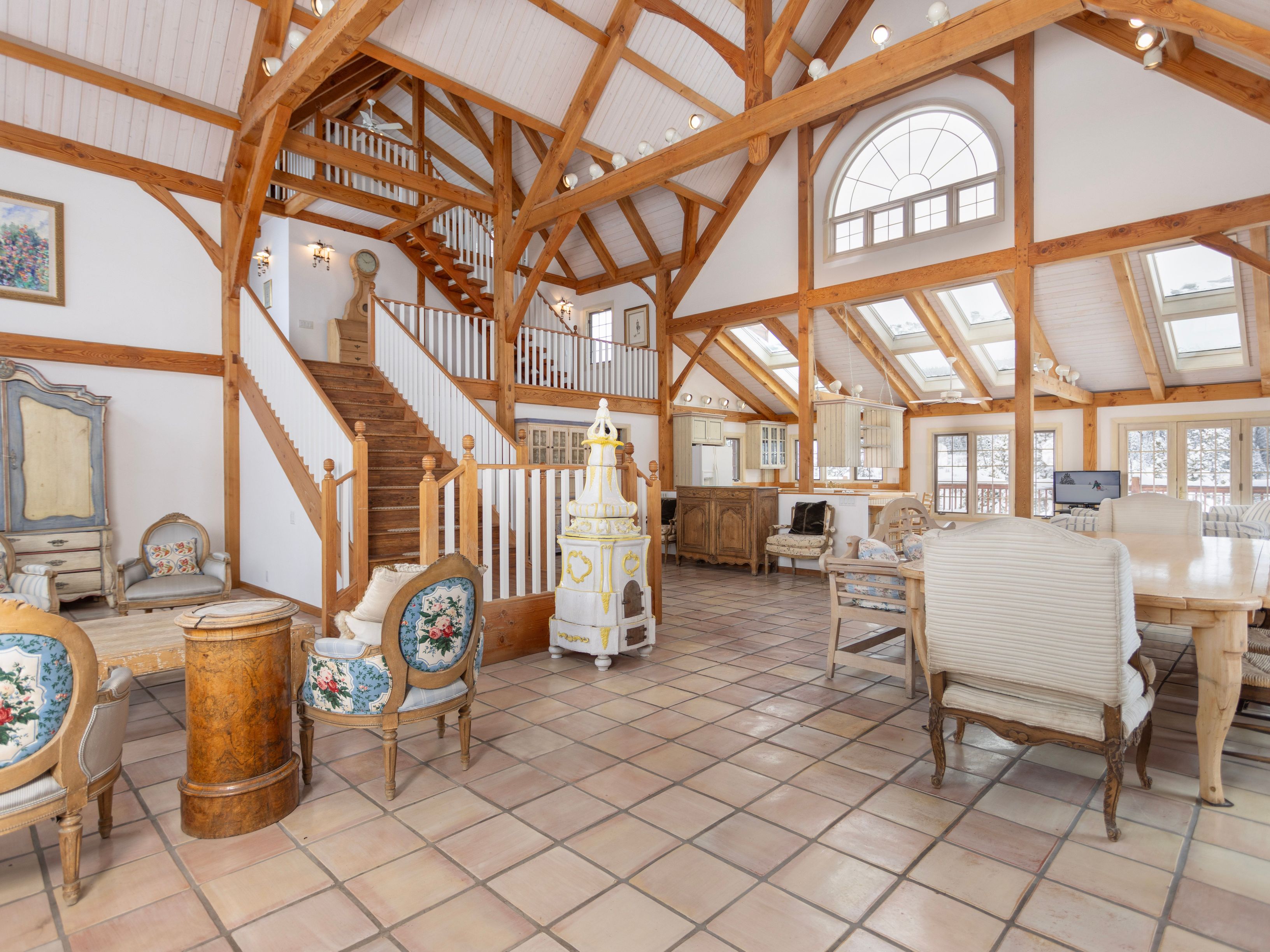This expansive, open-concept living and dining area features a stunning timber-frame construction with vaulted ceilings and exposed wooden beams. The space is anchored by a unique, ornate white and yellow ceramic stove, a grand staircase, and a mix of rustic and traditional furnishings set against light-colored tile flooring. Large windows and skylights flood the room with natural light, creating an airy, sophisticated atmosphere that blends architectural grandeur with cozy, classic comfort.