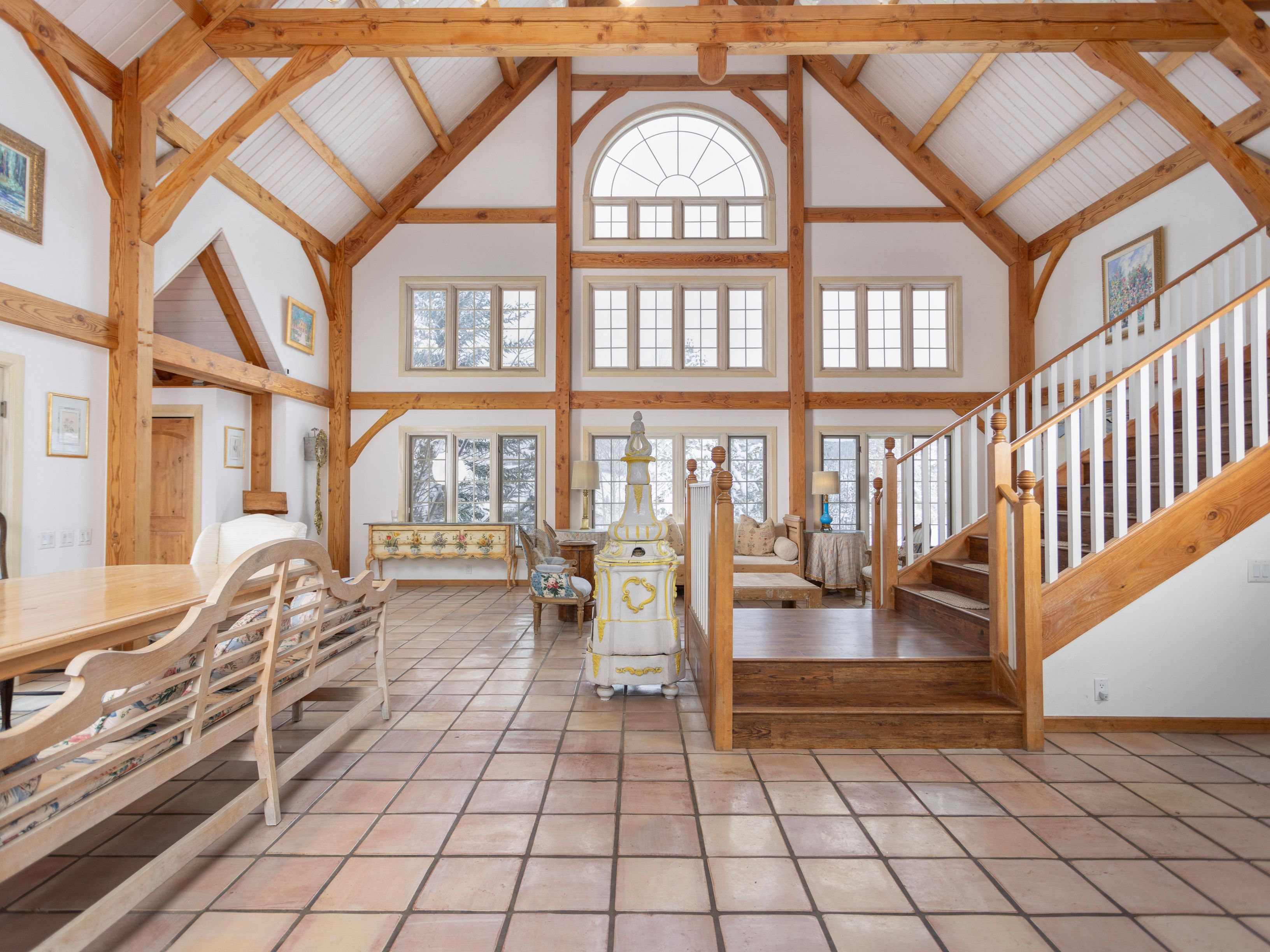 This expansive, open-concept living area features a dramatic vaulted ceiling with exposed timber beams and a striking floor-to-ceiling window wall that floods the space with natural light. The room is anchored by a unique, ornate ceramic stove and features rustic terracotta tile flooring, a wooden staircase with white railings, and a warm, traditional aesthetic. The perspective is centered, showcasing the grand scale of the architecture and the seamless transition between the dining and lounge areas.