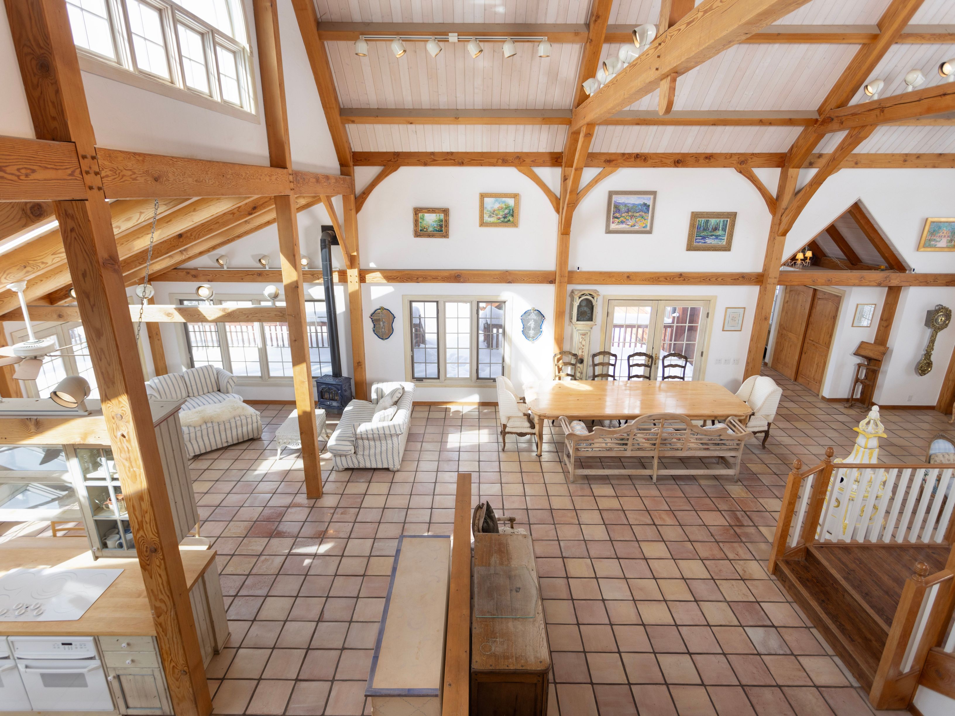 This expansive, open-concept living and dining area features a dramatic vaulted ceiling with exposed timber beams and a rustic, airy aesthetic. The space is anchored by a cozy wood-burning stove, a large dining table, and tiled flooring, all bathed in natural light from high windows and glass doors. The elevated perspective captures the grand scale of the room, highlighting its blend of traditional craftsmanship and comfortable, inviting living space.