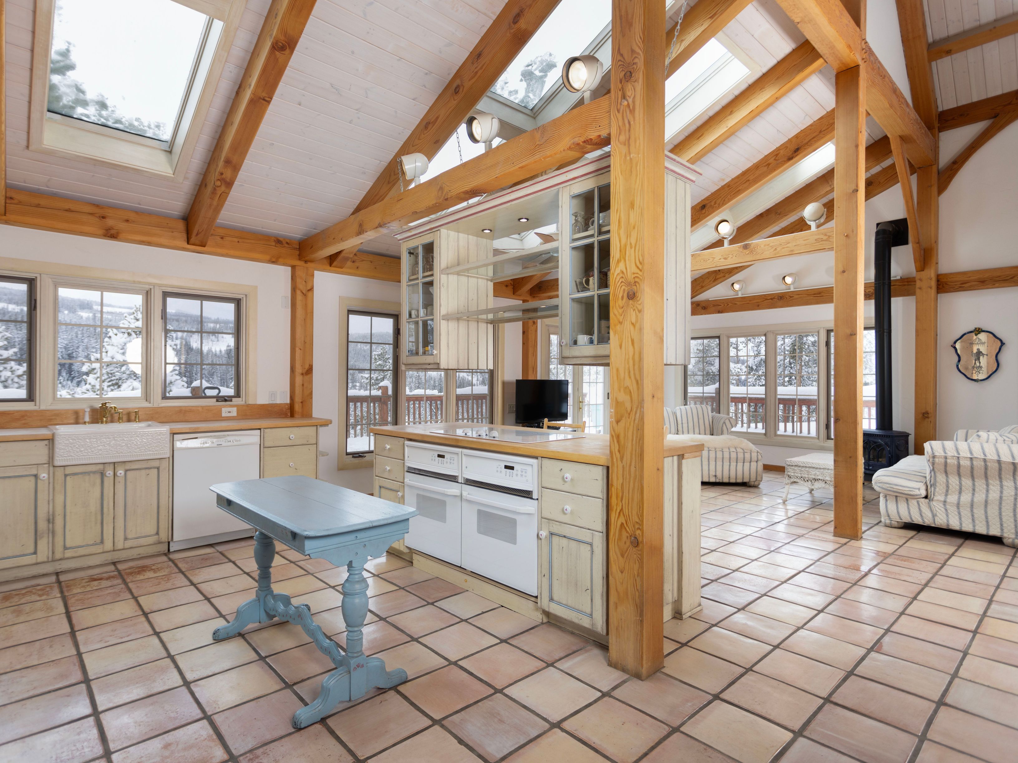 This rustic, open-concept kitchen features exposed timber beams, vaulted ceilings with skylights, and a unique central island with a built-in oven and cabinetry. The space is anchored by a light blue antique-style table and terracotta tile flooring, creating a warm, cabin-like atmosphere. Large windows provide expansive views of a snowy landscape, seamlessly blending the cozy interior with the natural beauty outside.