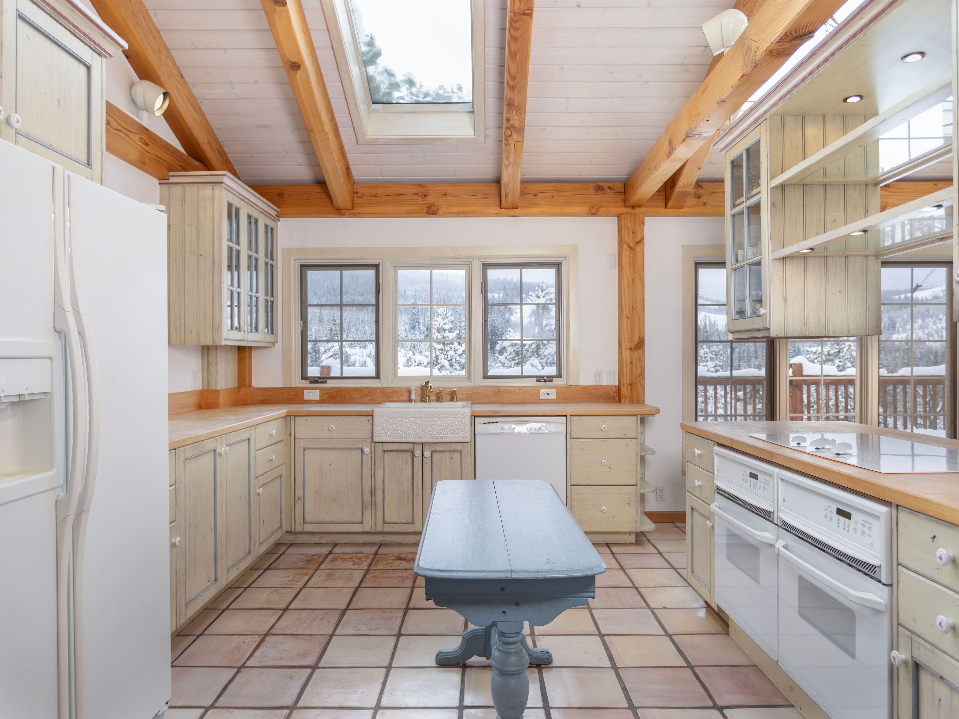 This rustic-style kitchen features light-toned wooden cabinetry, a farmhouse sink, and a central blue wooden island, all set against a backdrop of exposed timber beams and a vaulted ceiling with a skylight. The space is brightened by large windows that offer a scenic view of a snow-covered landscape, creating a cozy, mountain-lodge atmosphere. The kitchen is equipped with white appliances and finished with warm-toned tile flooring, blending traditional charm with functional design.