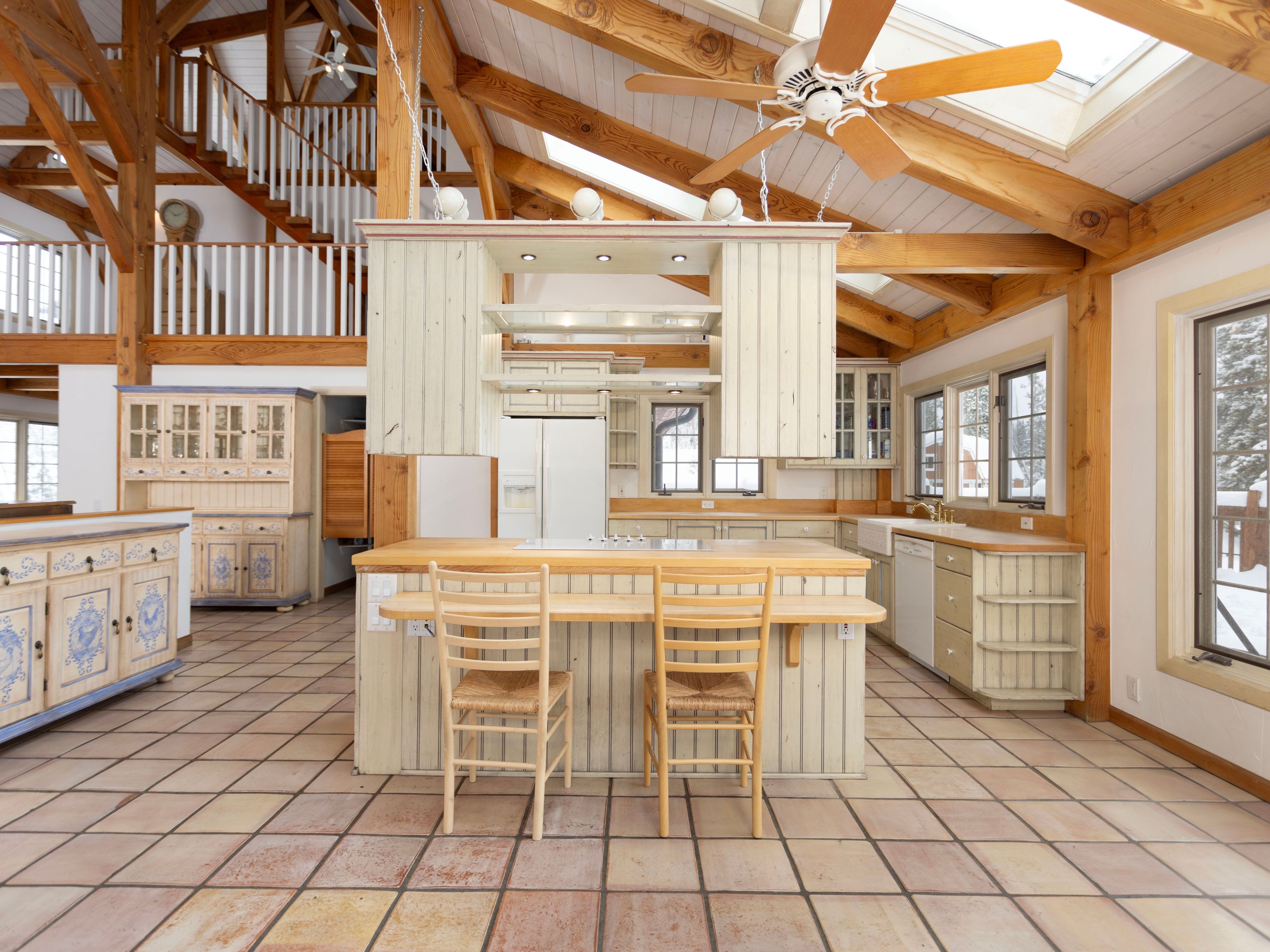 This spacious, rustic-style kitchen features a large central island with a wooden countertop and two chairs, positioned beneath a unique hanging cabinet structure. The room is defined by its impressive vaulted timber-frame ceiling, exposed beams, and warm, earthy tile flooring, creating a cozy yet expansive atmosphere. A lofted walkway overlooks the kitchen, adding architectural depth and a sense of openness to this charming, country-inspired home.