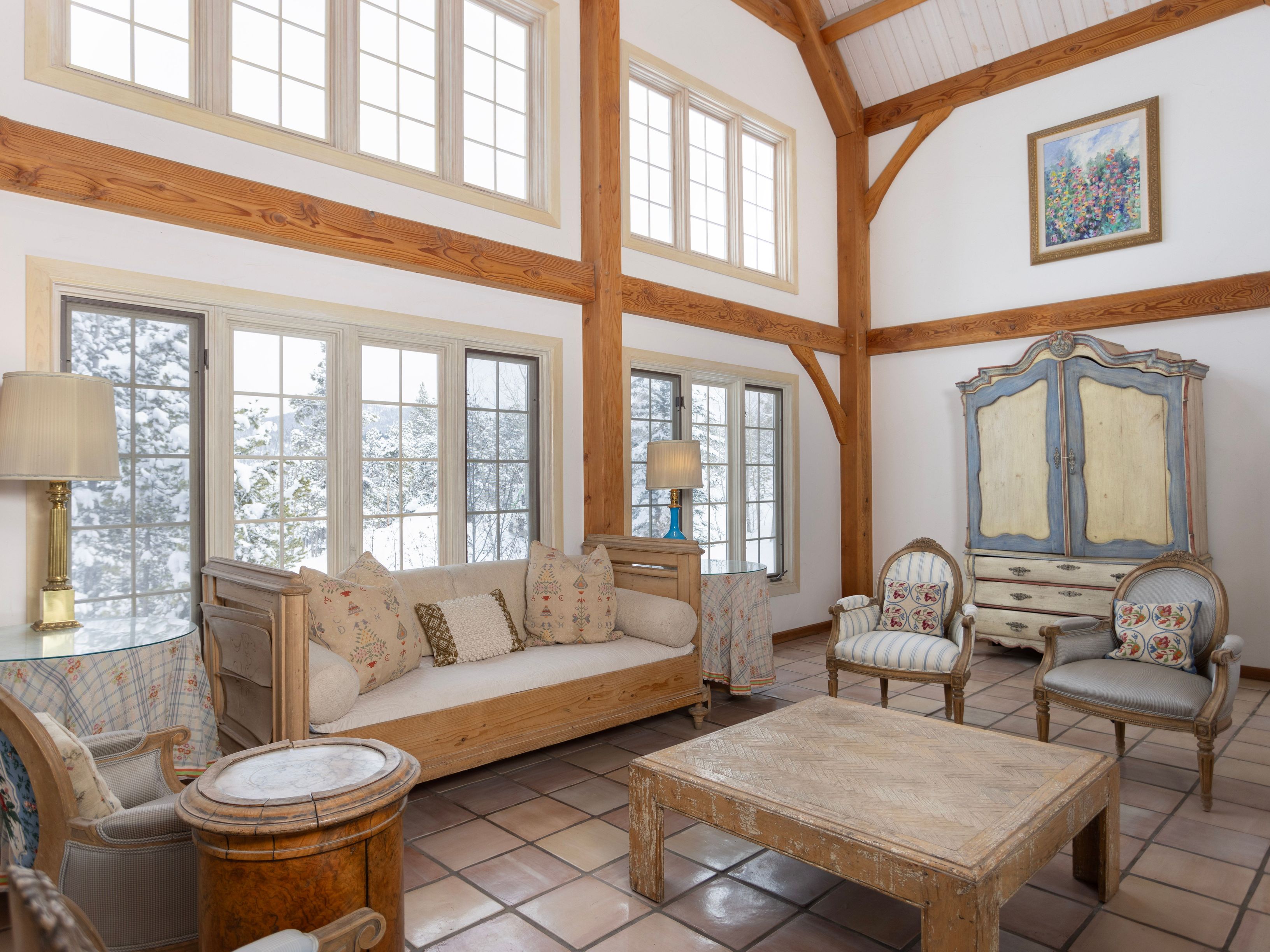 This spacious, light-filled living room features a rustic timber-frame design with soaring vaulted ceilings and expansive windows that offer a serene view of a snowy landscape. The room is furnished with a classic wooden daybed, elegant upholstered armchairs, and a large, weathered coffee table, all set against a backdrop of warm terracotta tile flooring. A decorative painted armoire and soft, patterned textiles add a touch of traditional charm to this cozy, mountain-inspired retreat.