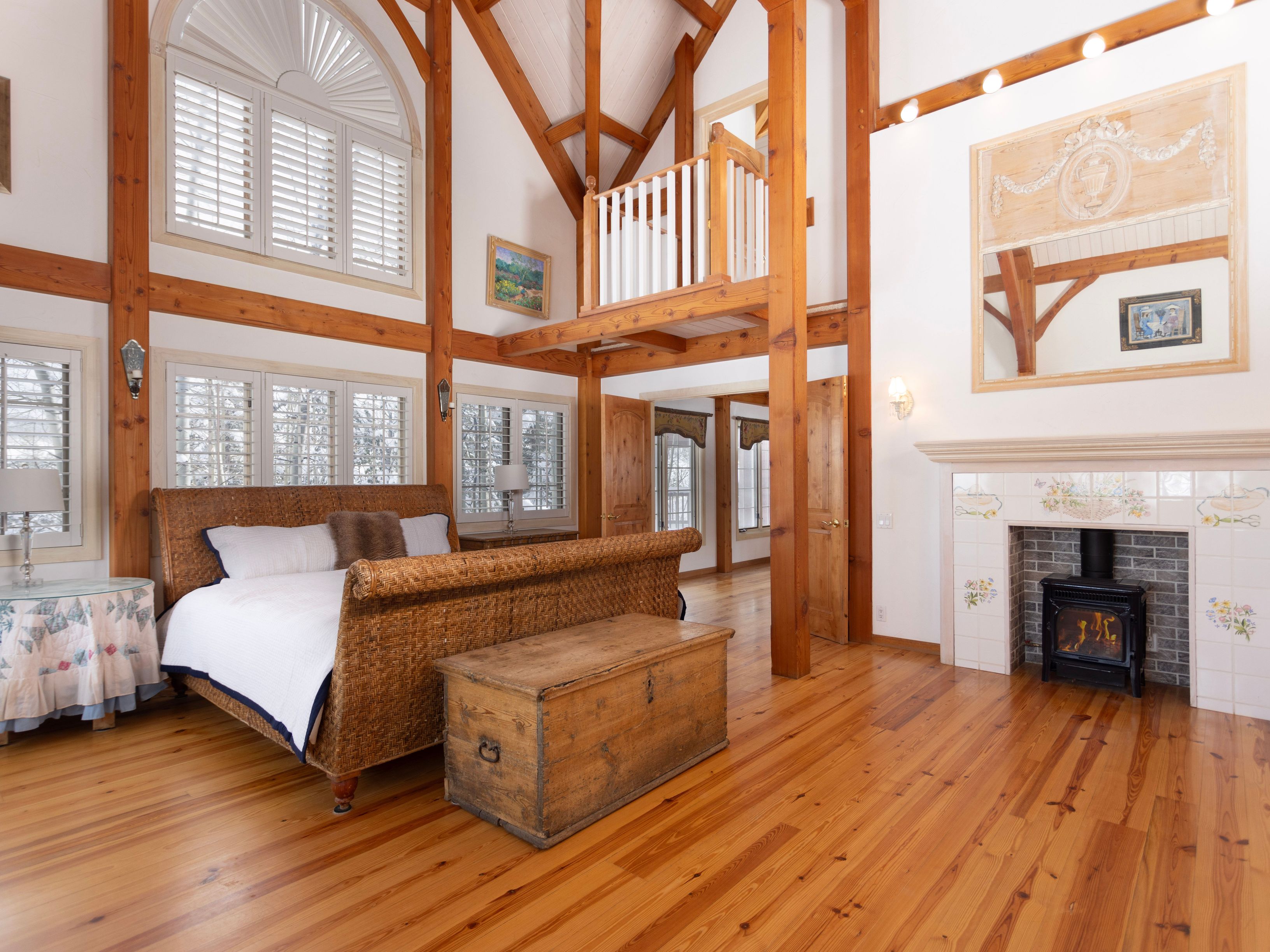 This spacious primary bedroom features a rustic, timber-frame architectural style with high vaulted ceilings and exposed wooden beams. The room is anchored by a large wicker sleigh bed, a vintage wooden storage chest, and a cozy fireplace with decorative floral tiles. A lofted balcony overlooks the space, adding architectural interest and a sense of grandeur to this warm, inviting retreat.