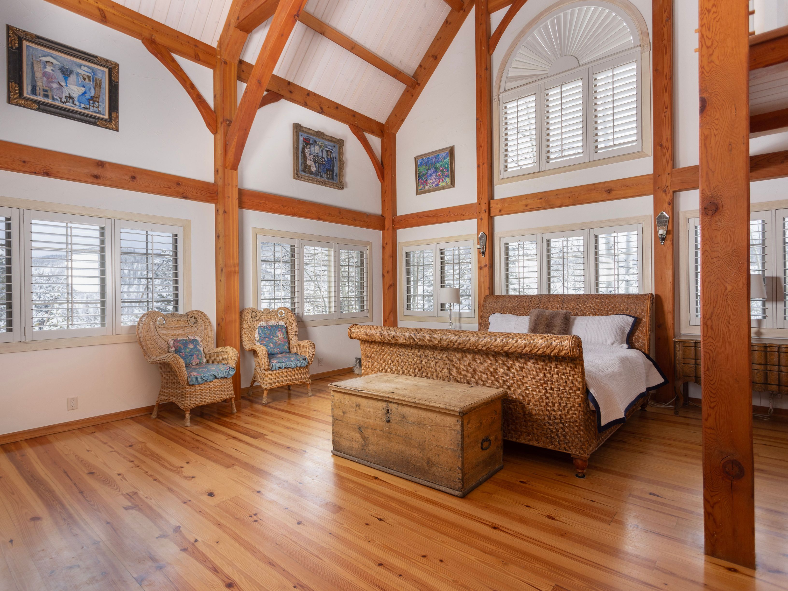 This spacious primary bedroom features a rustic, timber-frame architectural style with high vaulted ceilings and exposed wooden beams. The room is anchored by a large wicker sleigh bed and a vintage wooden trunk, complemented by two matching wicker armchairs and warm hardwood flooring. Large windows with white plantation shutters allow natural light to fill the space, creating an airy and serene atmosphere.