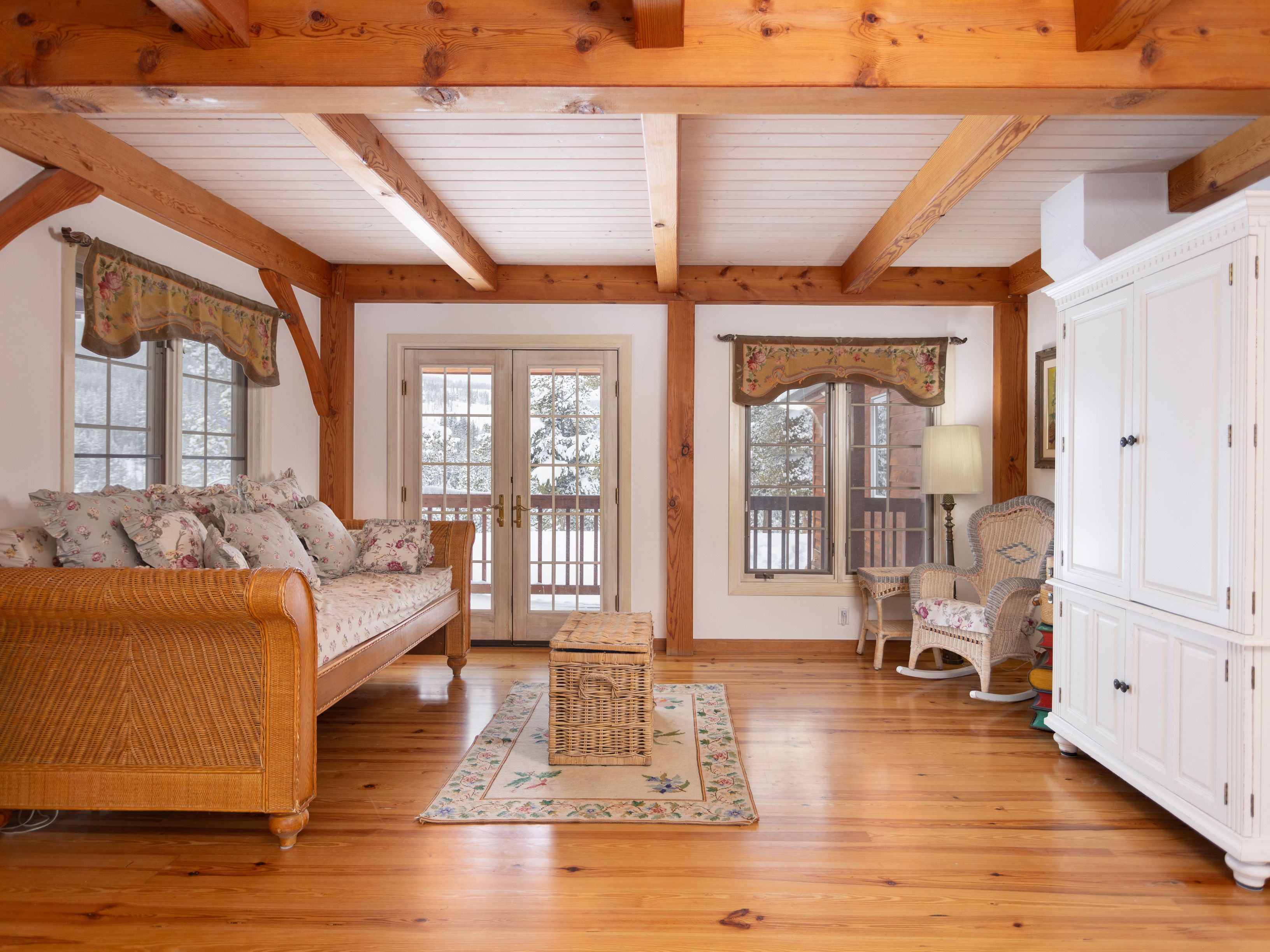 This cozy, rustic living room features exposed wooden ceiling beams, warm hardwood flooring, and a large wicker daybed adorned with floral cushions. A central wicker trunk sits on a decorative area rug, while a white armoire and a matching wicker rocking chair complete the charming, cottage-inspired aesthetic. The room is bathed in natural light from the glass-paned double doors and windows, which offer a serene view of a snowy landscape.