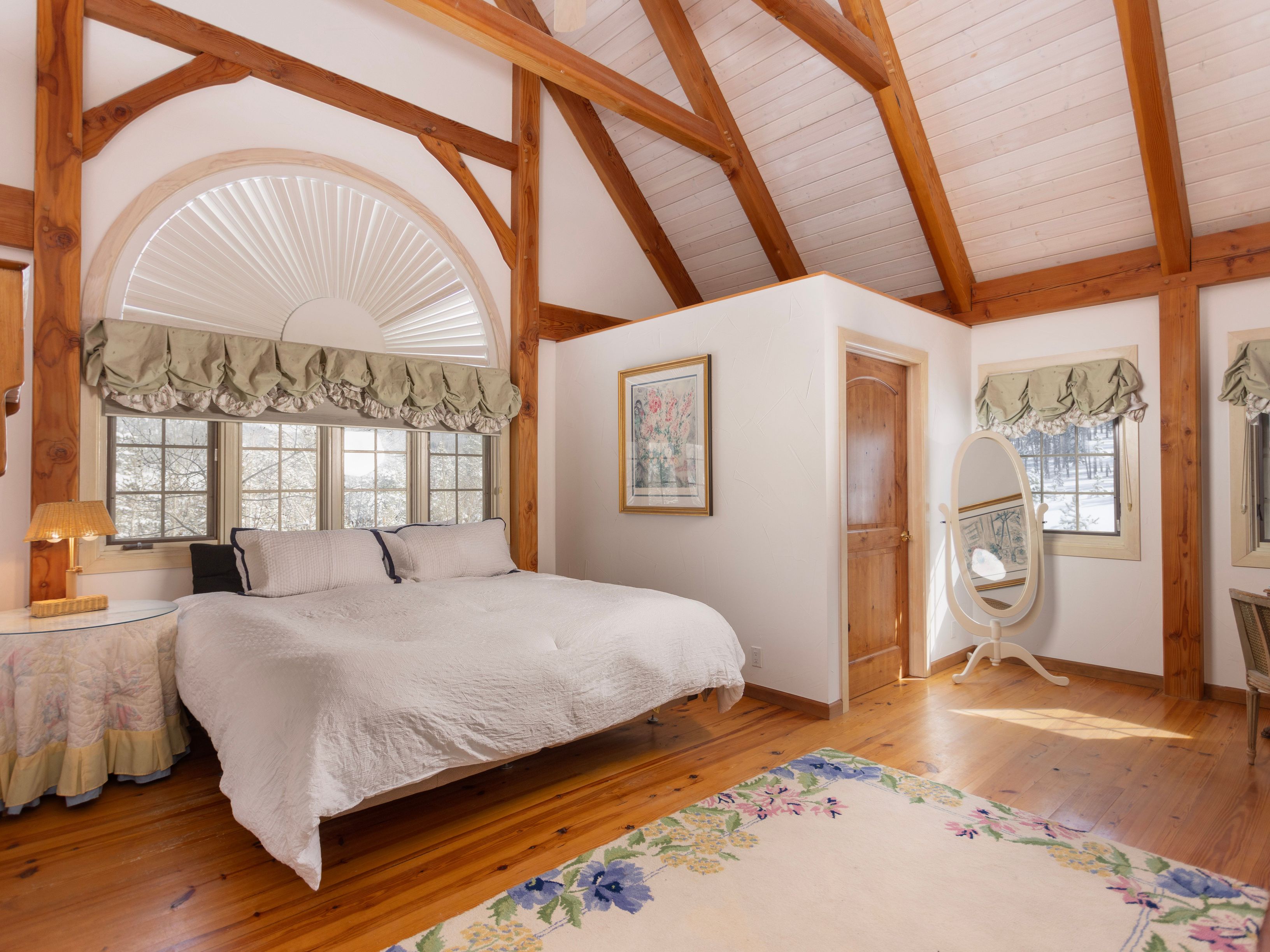 This spacious primary bedroom features a rustic aesthetic with exposed wooden beams and a vaulted ceiling. A large, arched window with a pleated shade serves as a focal point above the bed, while a floral area rug adds a touch of softness to the warm-toned hardwood floors. The room includes a standalone oval mirror and a private door, creating a cozy and inviting atmosphere.