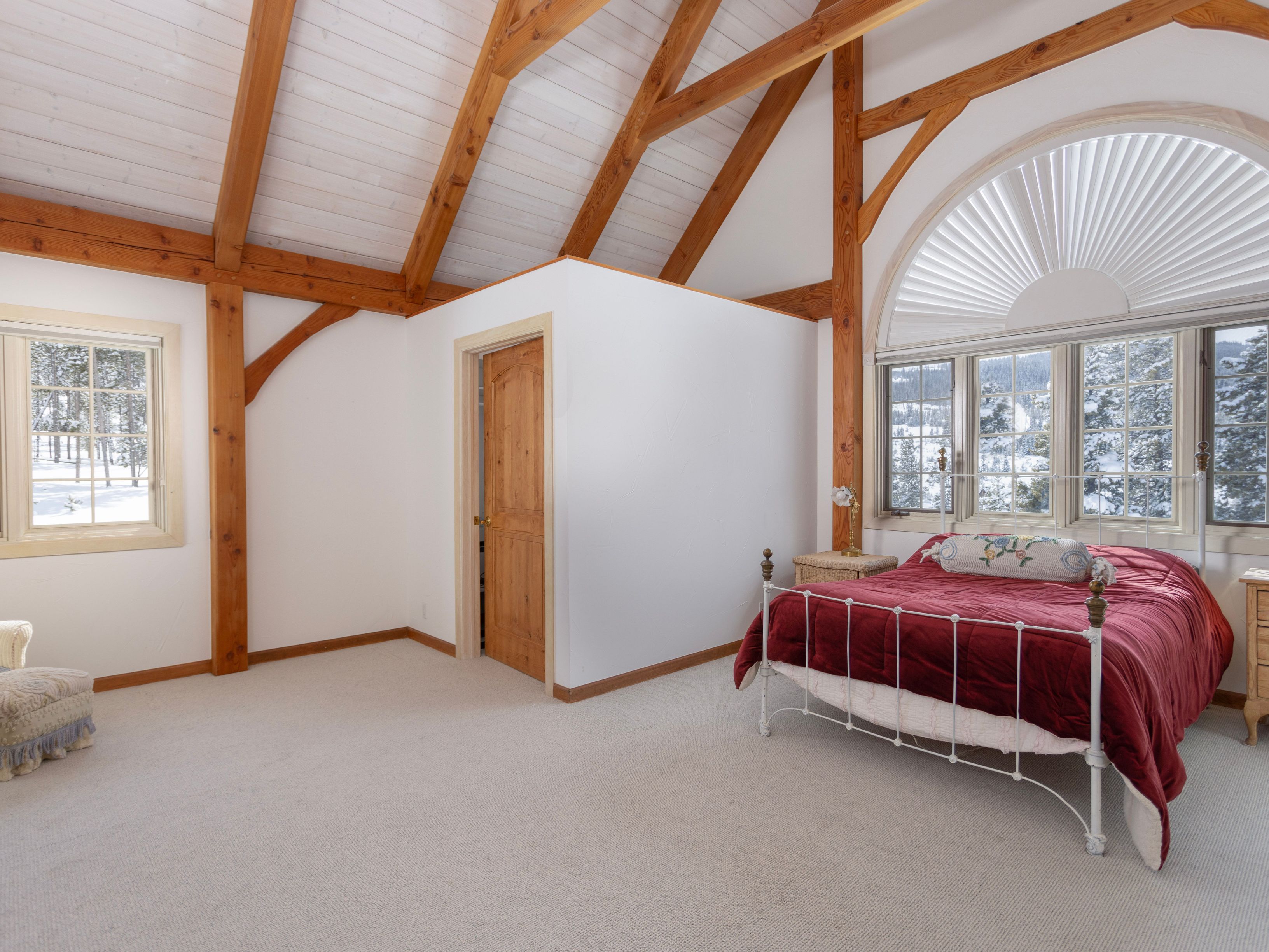 This spacious bedroom features a rustic aesthetic with exposed timber beams and a vaulted ceiling, creating an airy and inviting atmosphere. The room is anchored by a white metal-framed bed with a vibrant red quilt and a large, arched window that offers a scenic view of a snowy landscape. A neutral carpet and a simple wooden door leading to a closet complete the functional and cozy design.