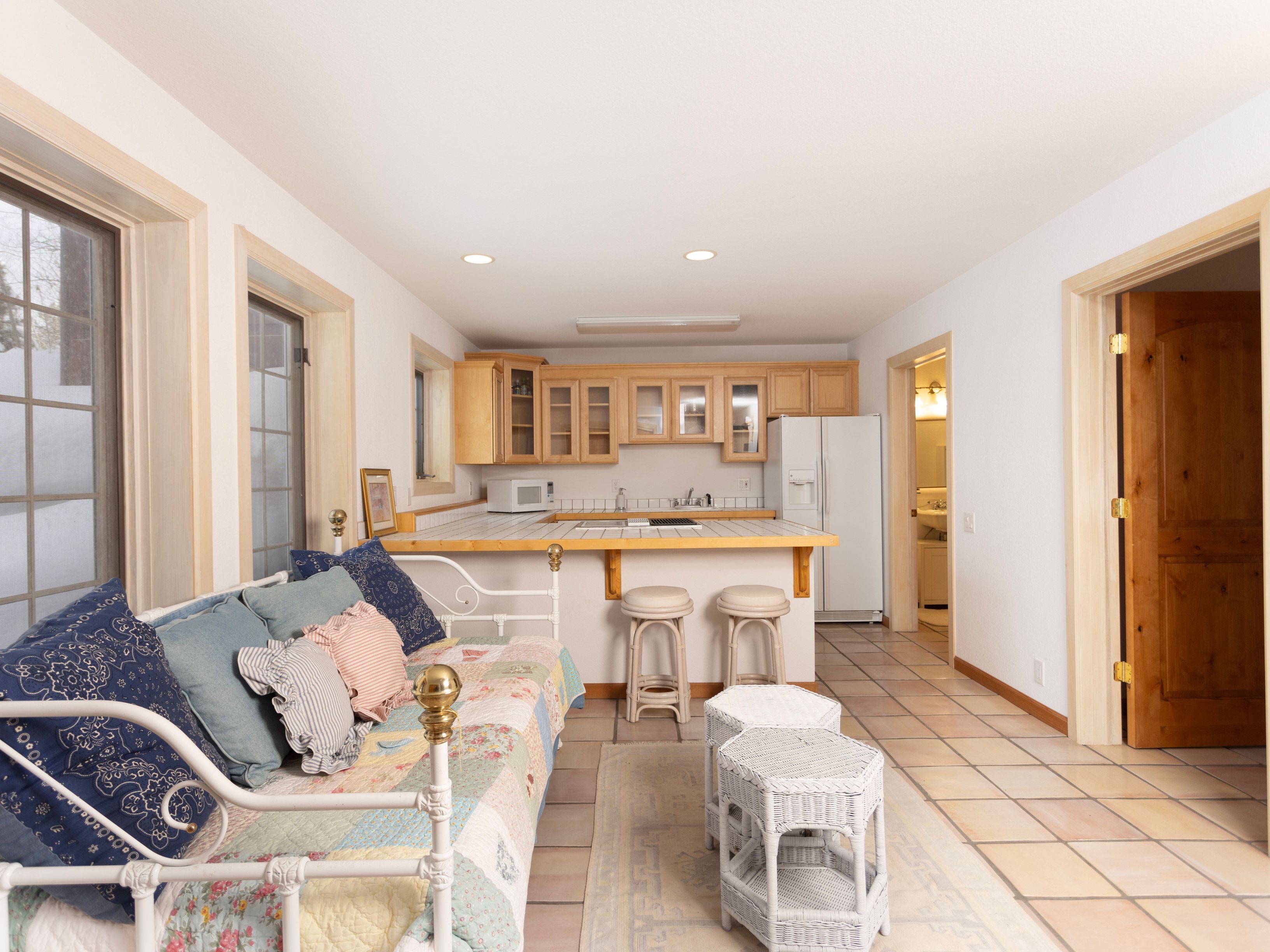 This open-concept space features a cozy seating area with a daybed in the foreground, transitioning into a functional kitchen with light wood cabinetry and tiled countertops. The room is bright and inviting, with a neutral color palette, tiled flooring, and a clear view into an adjacent bathroom. The perspective is captured from a low angle, emphasizing the seamless flow between the living and kitchen areas.