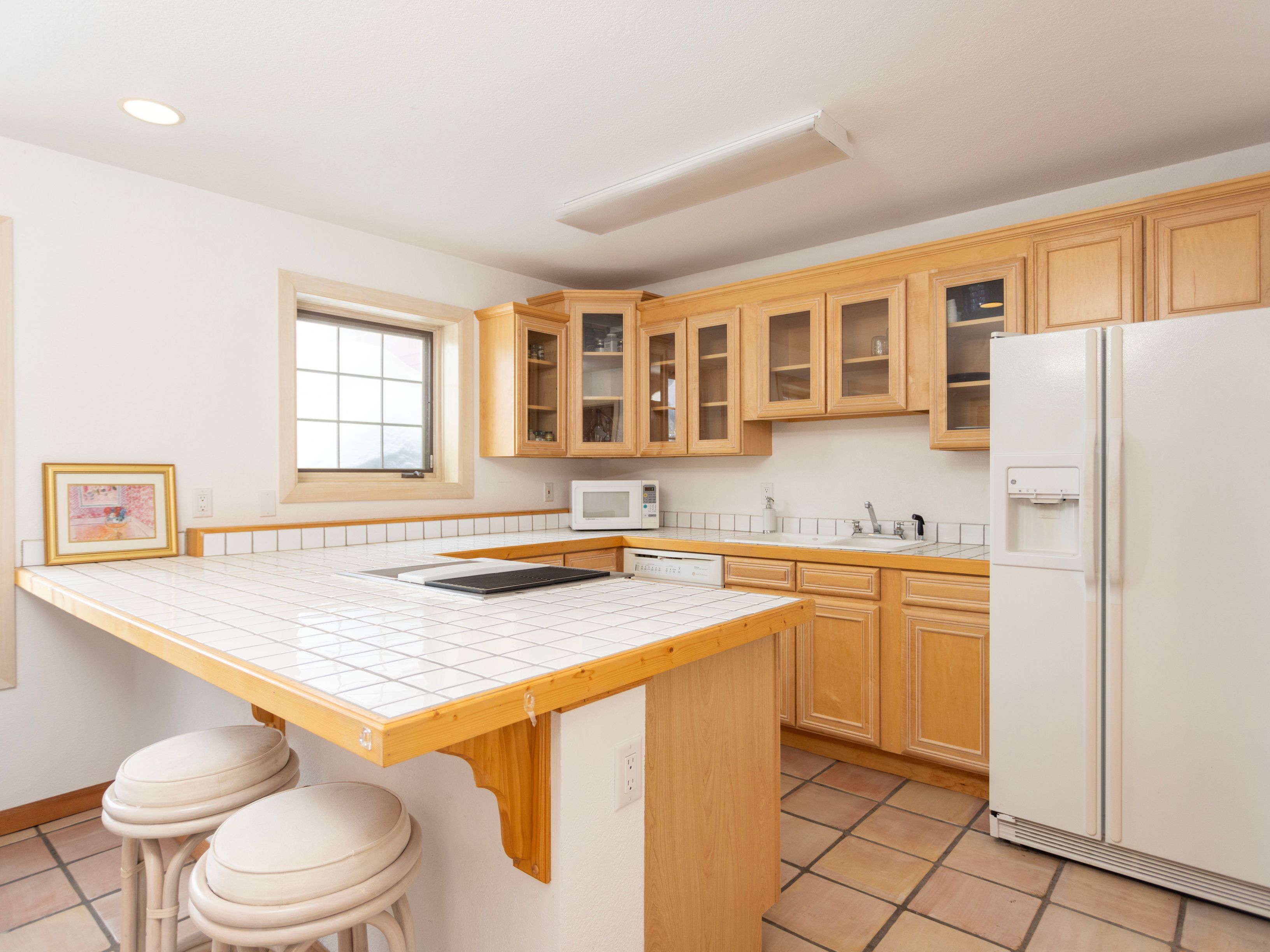 This kitchen features a U-shaped layout with light wood cabinetry, white tiled countertops, and a matching tiled breakfast bar with two stools. The space includes a white refrigerator, a built-in microwave, and a sink area, all set against a backdrop of terracotta-style floor tiles. The perspective is a wide-angle shot from the breakfast bar, showcasing the functional workspace and the bright, neutral aesthetic of the room.