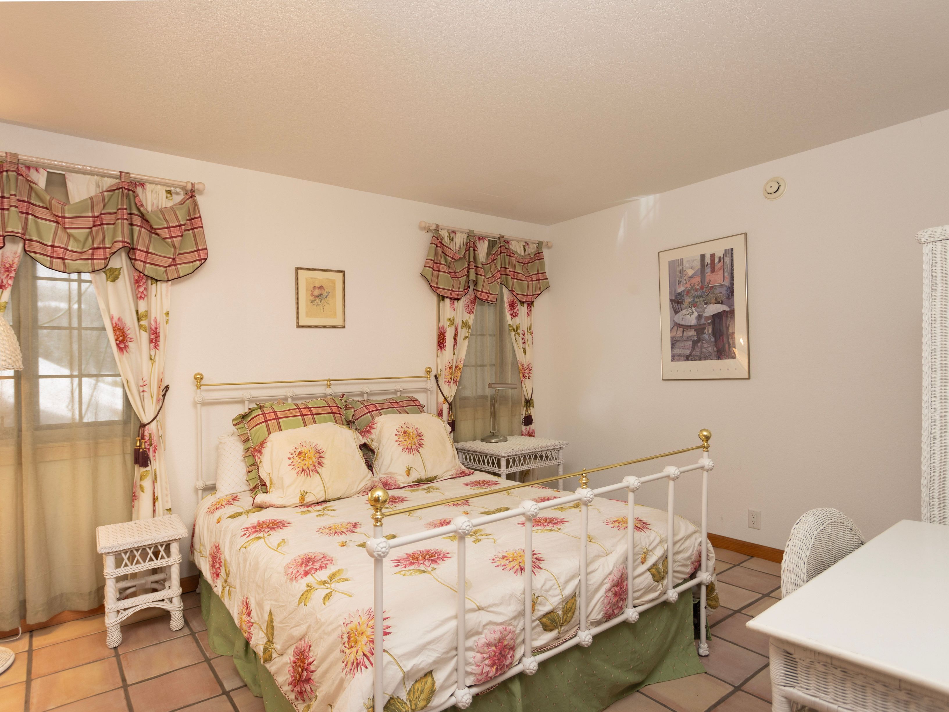 This cozy guest bedroom features a classic white metal bed frame adorned with floral bedding and matching plaid pillows. The room is accented by two white wicker nightstands, floral curtains with valances, and tiled flooring, creating a charming, traditional aesthetic. The perspective is a wide-angle shot from the corner of the room, capturing the bed as the central focal point.
