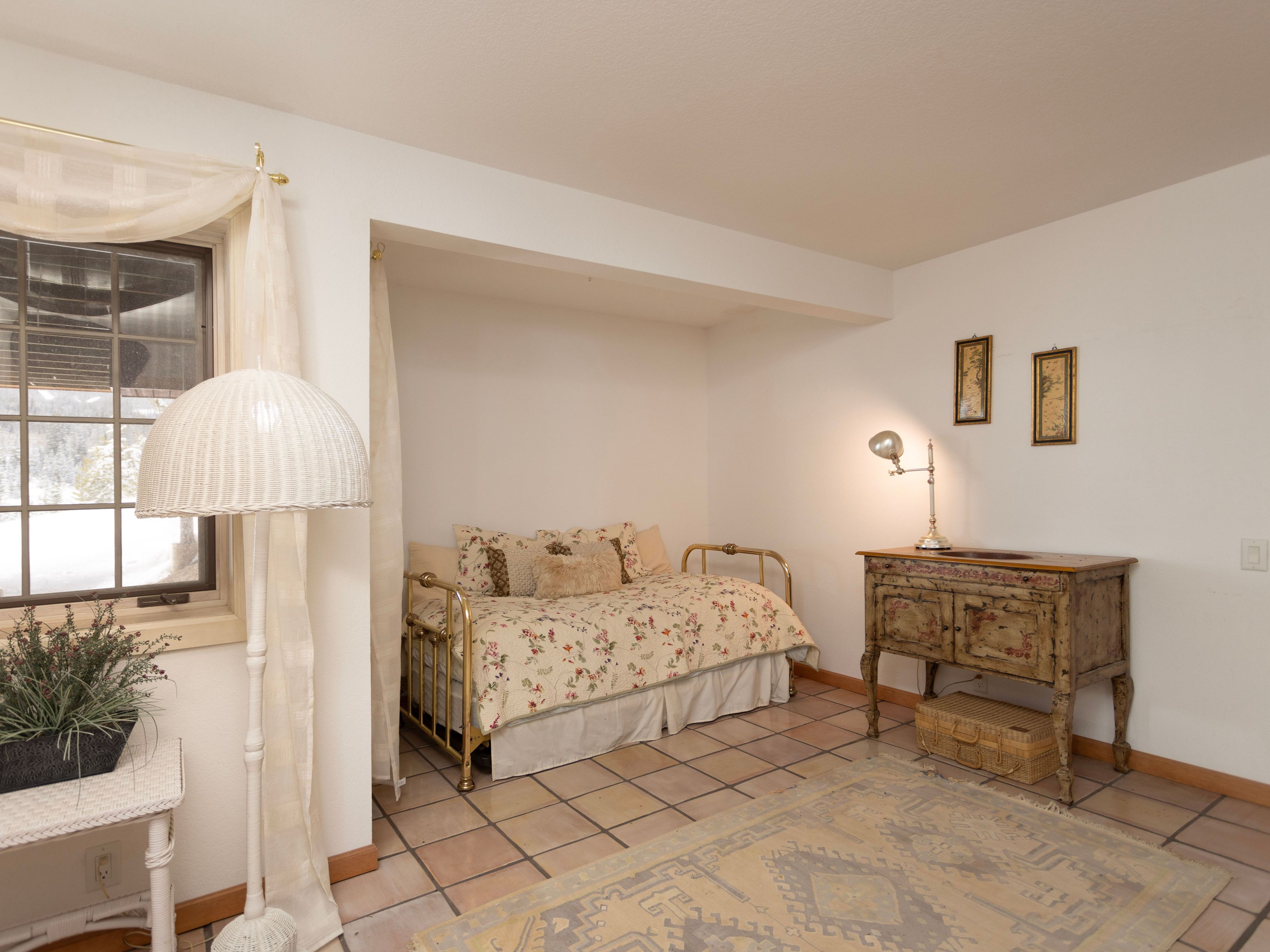 This cozy guest bedroom features a classic brass daybed adorned with a floral quilt and decorative pillows, set within a recessed alcove. The room is accented by a vintage-style wooden vanity with a lamp, a patterned area rug over tile flooring, and a white wicker floor lamp near the window. The overall aesthetic is charming and rustic, creating a warm and inviting atmosphere.