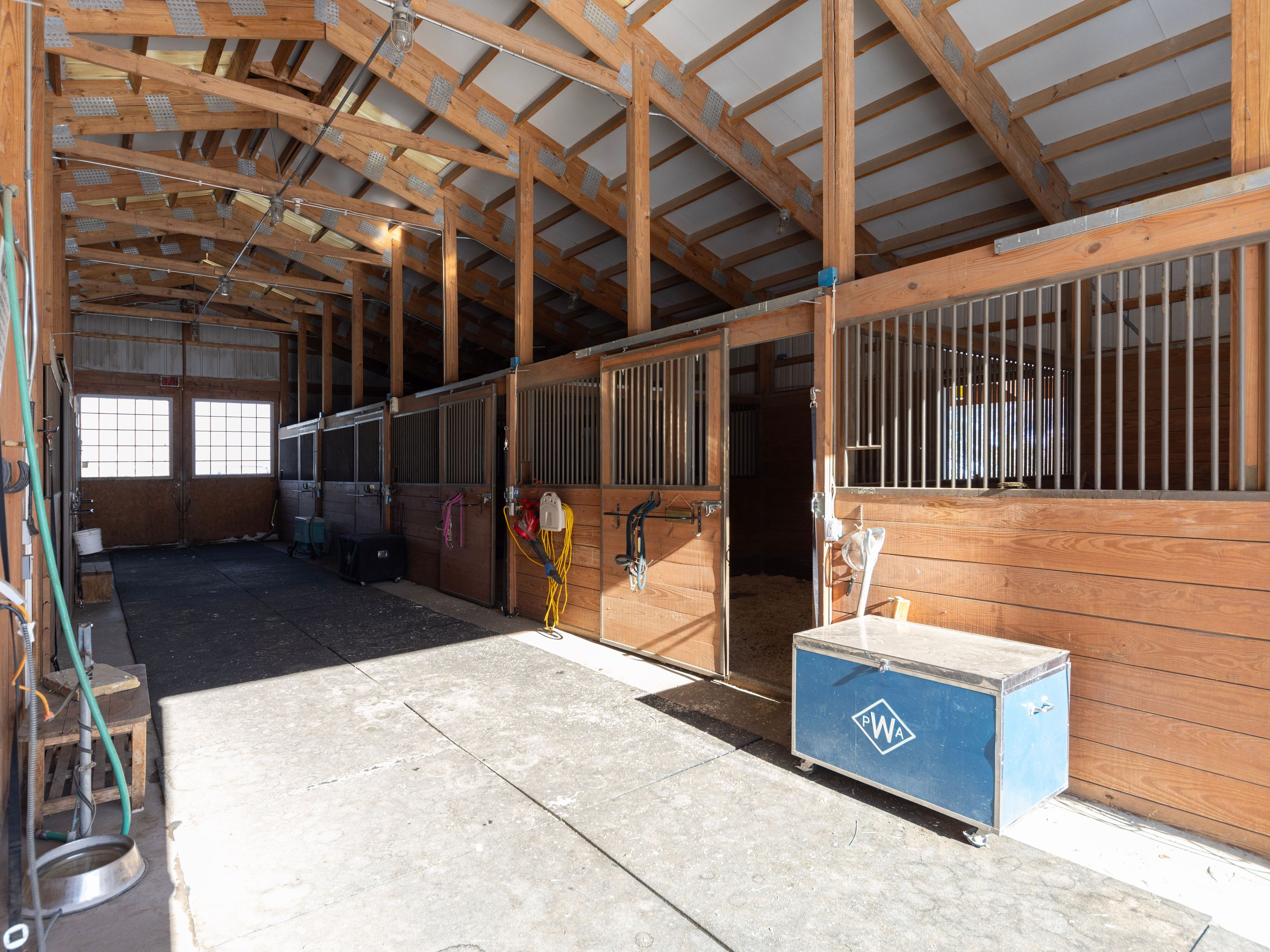This image captures the interior of a well-maintained horse stable, featuring a long concrete aisle lined with multiple wooden stalls. The structure showcases exposed timber framing and high ceilings, with natural light streaming in from windows at the far end. A blue tack trunk sits in the foreground, and various equestrian equipment hangs on the stall doors, creating a functional and rustic atmosphere.