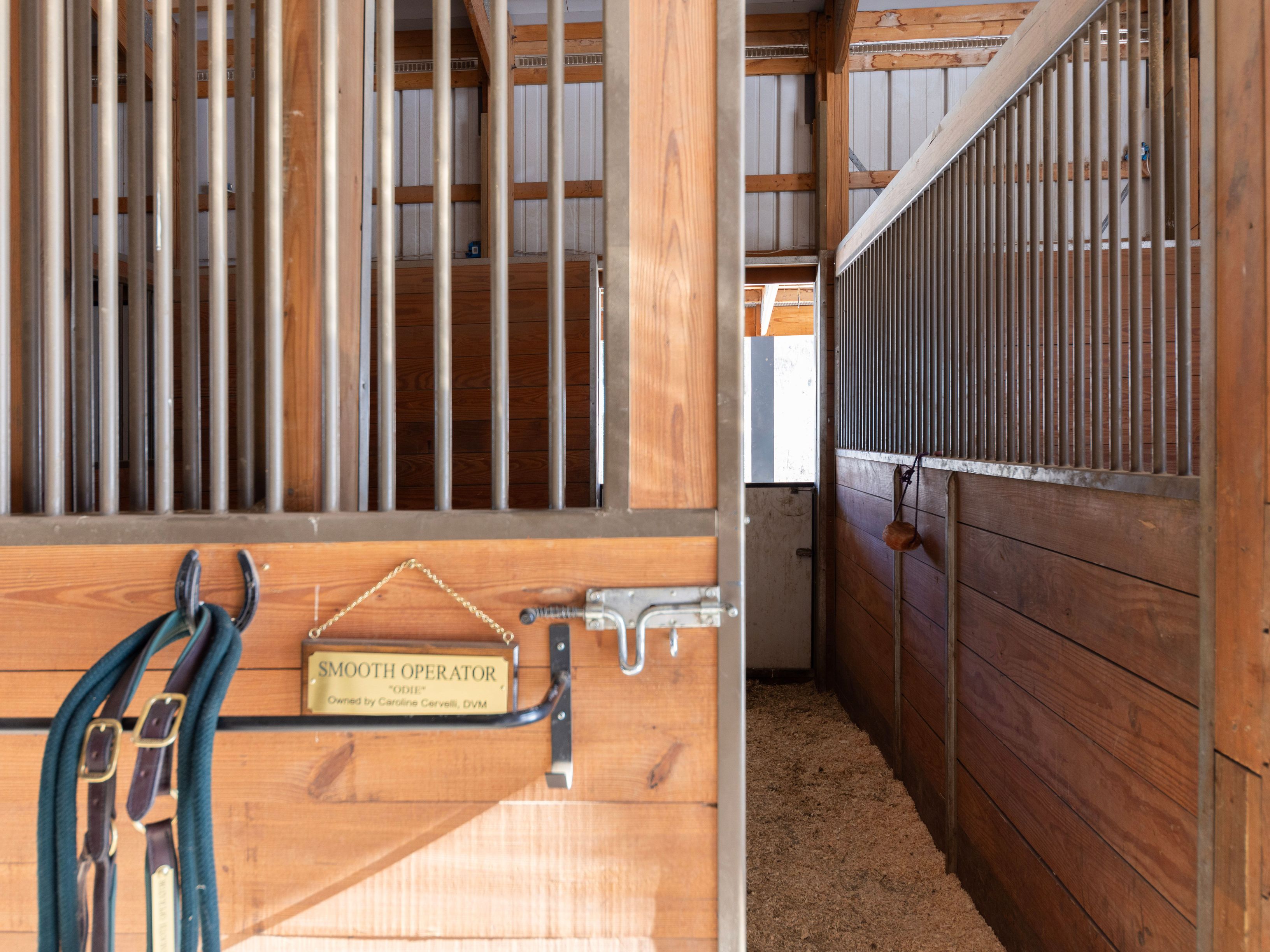 This image captures the interior of a well-maintained horse stable, featuring wooden stalls with metal bars and a nameplate for 'Smooth Operator' hanging on the gate. The perspective is from the aisle looking into a stall, highlighting the rustic wood construction and the clean, bedded floor. The scene conveys a professional and orderly equestrian environment.