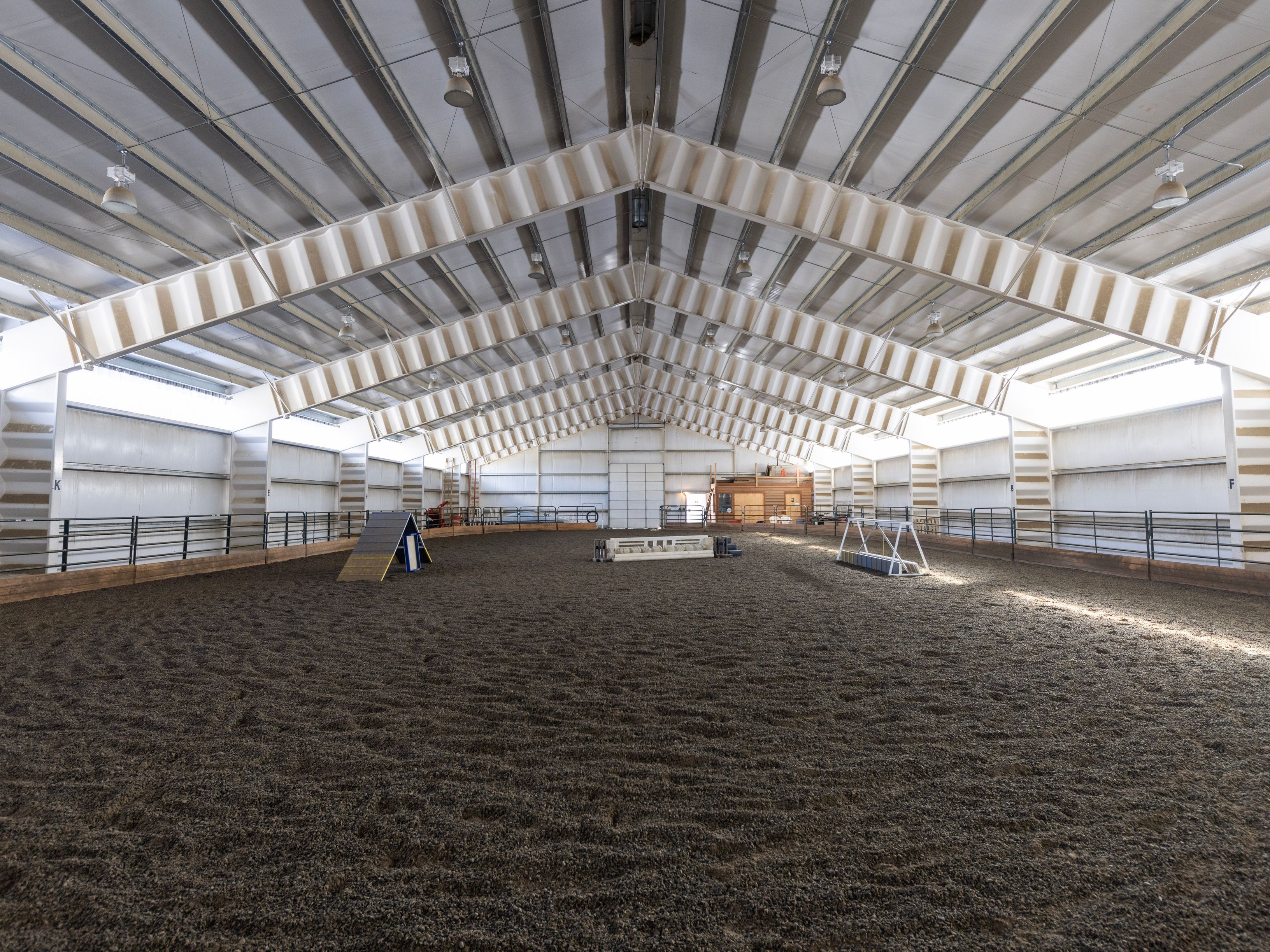 This image showcases a large, professional-grade indoor equestrian riding arena with a high-vaulted ceiling and exposed structural beams. The expansive space features a deep, well-maintained dirt footing, perimeter fencing, and various training obstacles, creating a functional environment for horse training. The perspective is a wide, centered shot that emphasizes the scale and open layout of the facility.