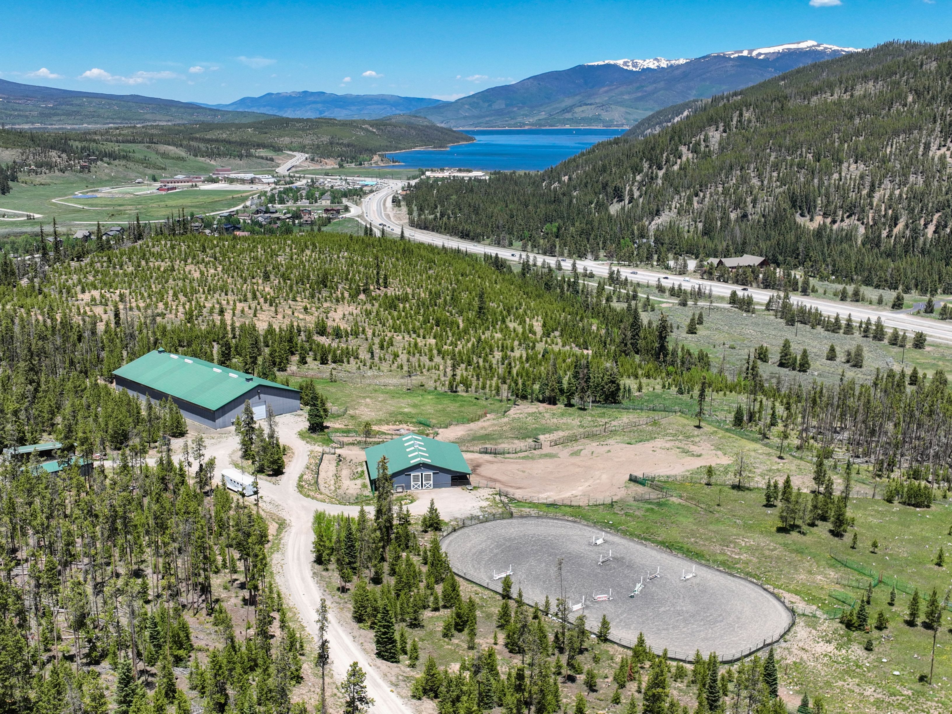 This high-angle aerial view captures a sprawling equestrian property nestled in a mountainous landscape, featuring two large green-roofed barns and a circular riding arena. The property is surrounded by dense pine forests and offers a stunning backdrop of a distant blue lake and snow-capped peaks. The scene conveys a sense of rural tranquility and expansive outdoor space, ideal for horse enthusiasts or those seeking a mountain retreat.