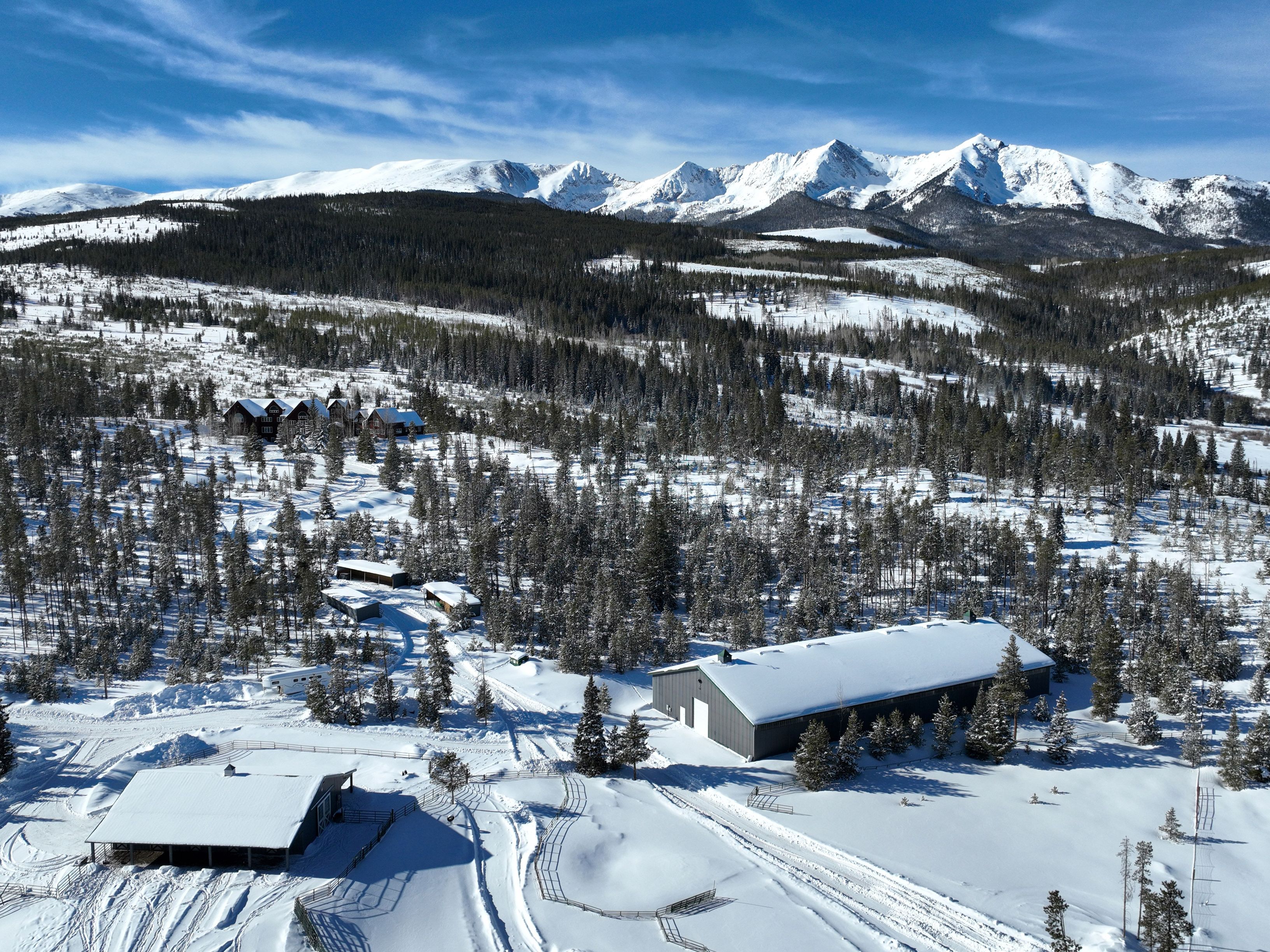 This expansive aerial view captures a sprawling mountain property blanketed in deep snow, featuring a large, modern metal-sided barn and a separate residential structure. The landscape is dotted with dense evergreen forests and connected by winding, snow-covered paths, all set against a dramatic backdrop of towering, snow-capped mountain peaks under a clear blue sky. The scene conveys a sense of serene, rugged isolation and high-altitude luxury.