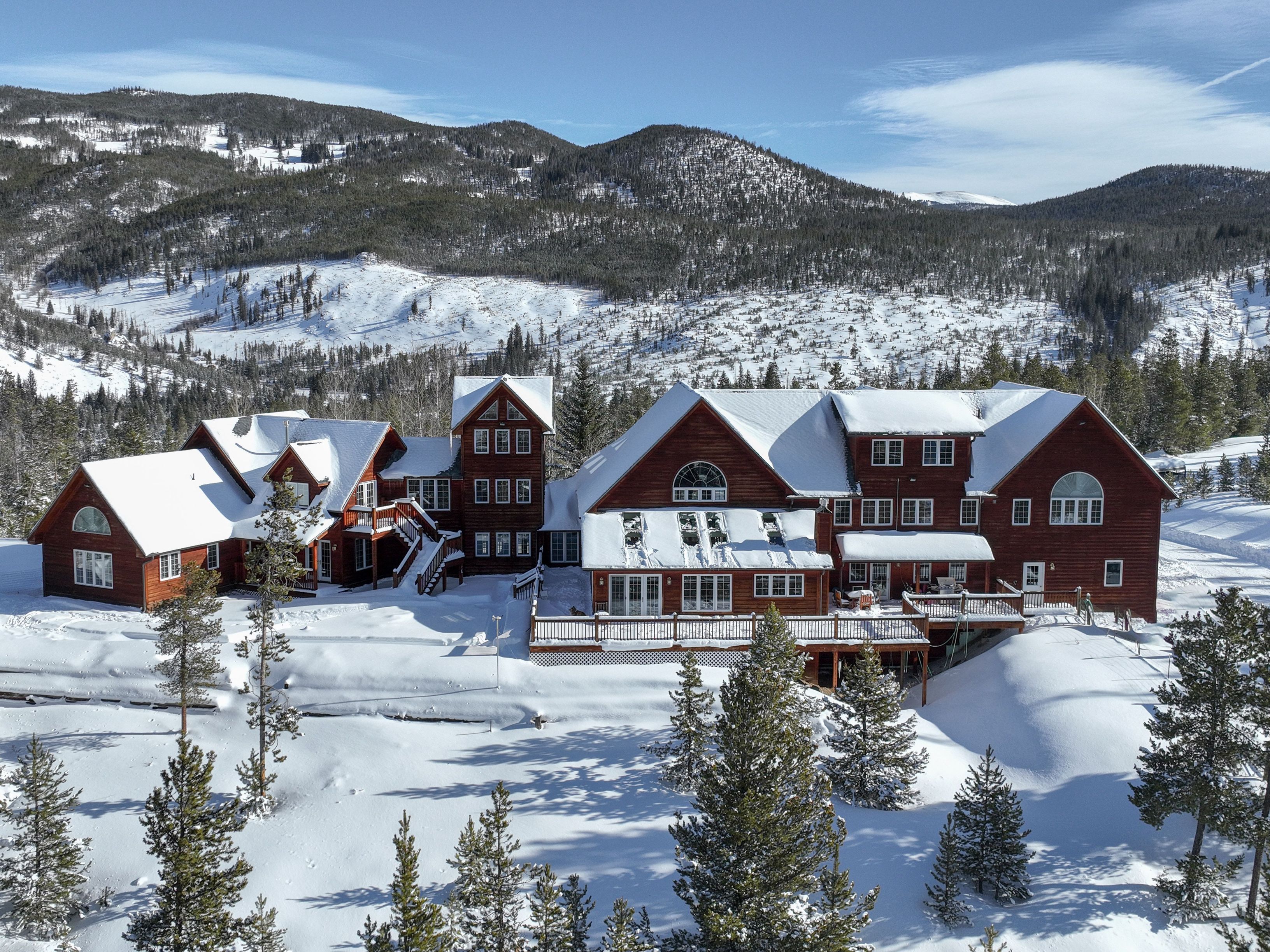 This stunning aerial shot captures a sprawling, multi-level log cabin estate nestled in a pristine, snow-covered mountain landscape. The architecture features classic rustic wood siding, numerous gabled rooflines, and expansive wooden decks that overlook the wintry forest and distant peaks. The bright, clear sky and deep snow create a serene, high-end mountain retreat atmosphere, perfect for a luxury winter getaway.