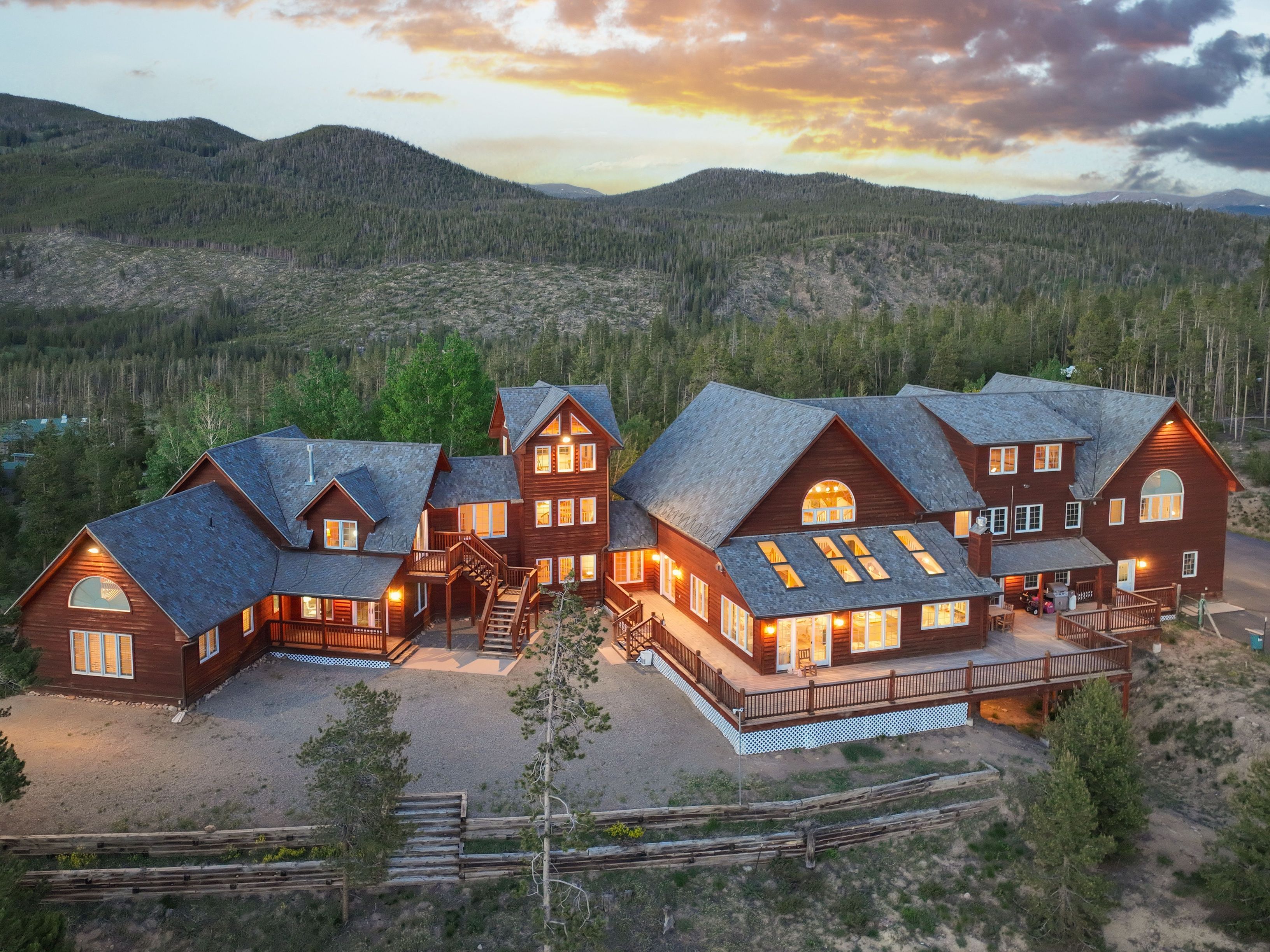 This stunning aerial perspective captures a sprawling, multi-level log cabin estate nestled against a backdrop of lush, forested mountains at sunset. The home features extensive wrap-around wooden decking, a prominent central tower, and a warm, inviting glow emanating from its many windows. The rustic architectural style blends seamlessly with the rugged natural landscape, creating an impressive and serene mountain retreat.