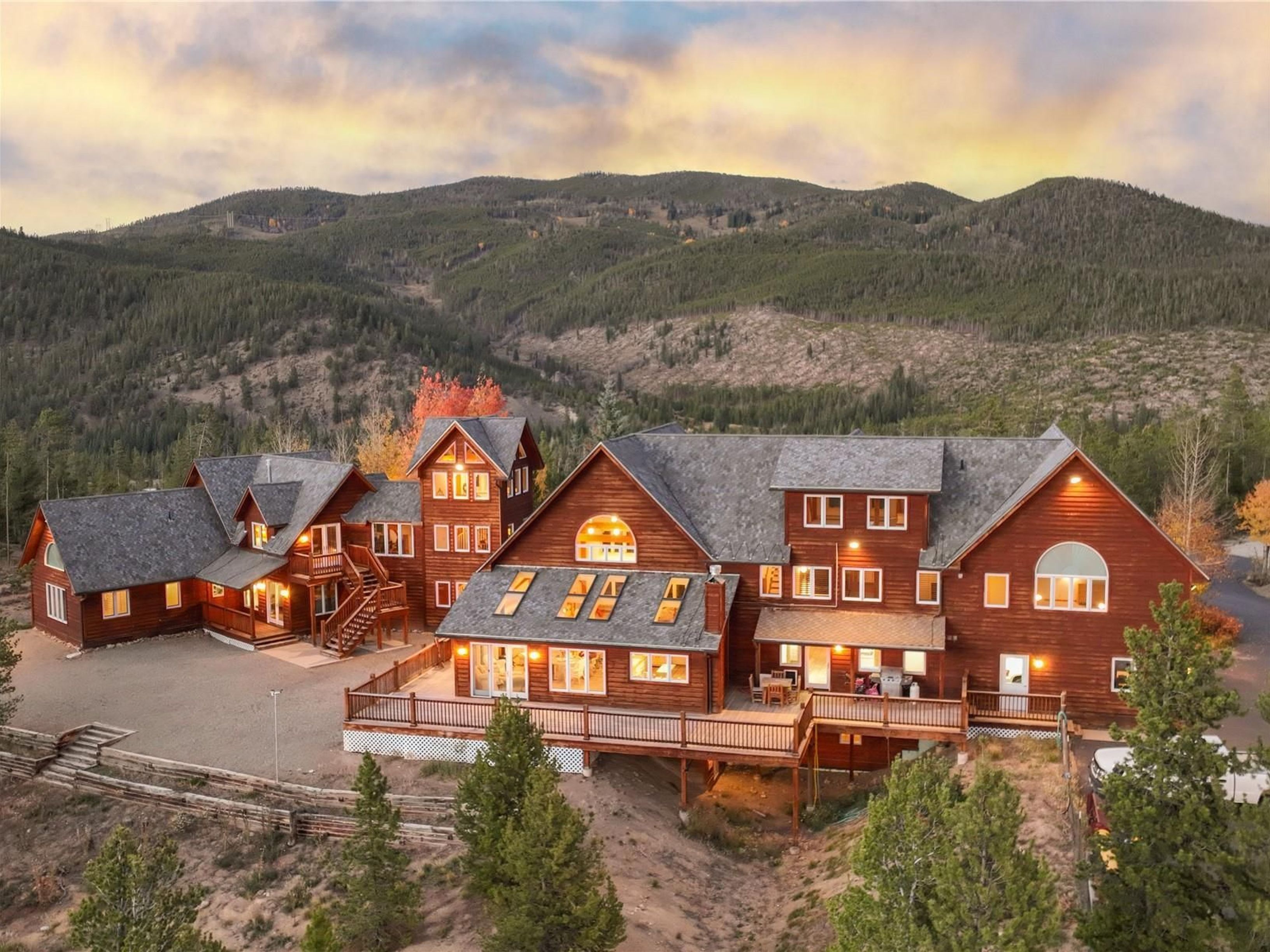 This expansive, multi-level mountain lodge features a classic log cabin aesthetic with warm wood siding, a dark shingled roof, and numerous windows that capture the surrounding landscape. The property includes a large, elevated wooden deck overlooking a rugged, forested hillside, creating a serene and secluded atmosphere. The aerial perspective highlights the home's impressive scale and its integration into the picturesque, mountainous terrain during a soft, golden-hour sunset.