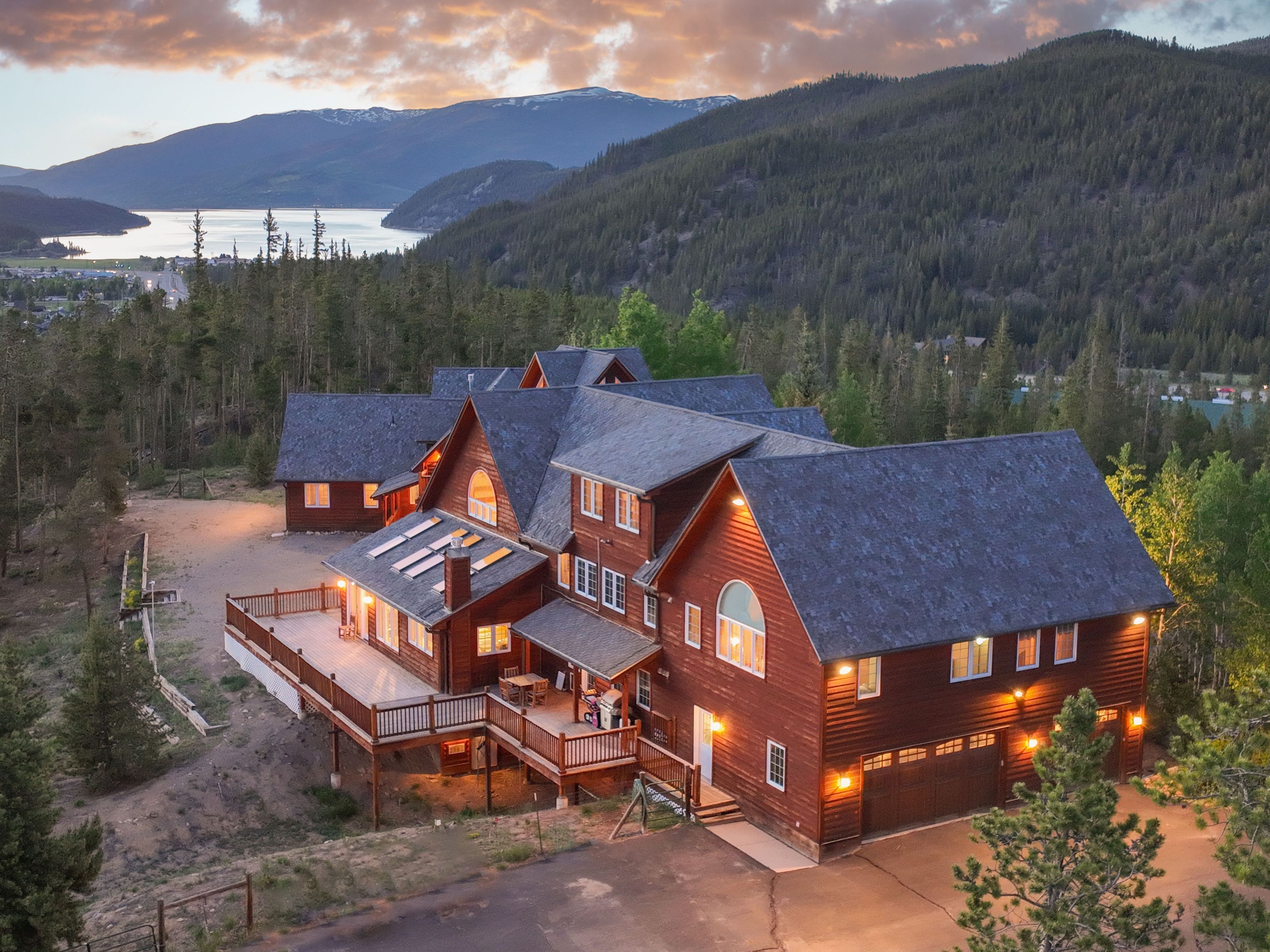This stunning aerial perspective captures a large, multi-level log cabin nestled into a forested mountainside at sunset. The home features a sprawling wooden deck, a multi-car garage, and a classic rustic aesthetic, all set against a breathtaking backdrop of a lake and distant snow-capped peaks. The warm glow of the interior and exterior lighting creates an inviting and luxurious atmosphere.