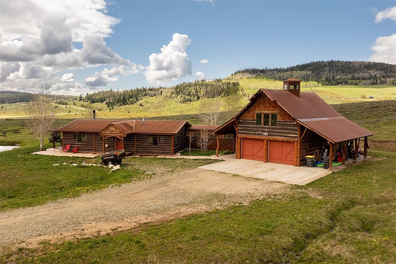 This aerial view showcases a charming log cabin and a barn-style garage, both featuring rustic wooden exteriors and reddish-brown roofs. The properties are set against a backdrop of rolling hills and lush greenery, creating a serene and picturesque setting. A gravel driveway connects the buildings, adding to the rural appeal.