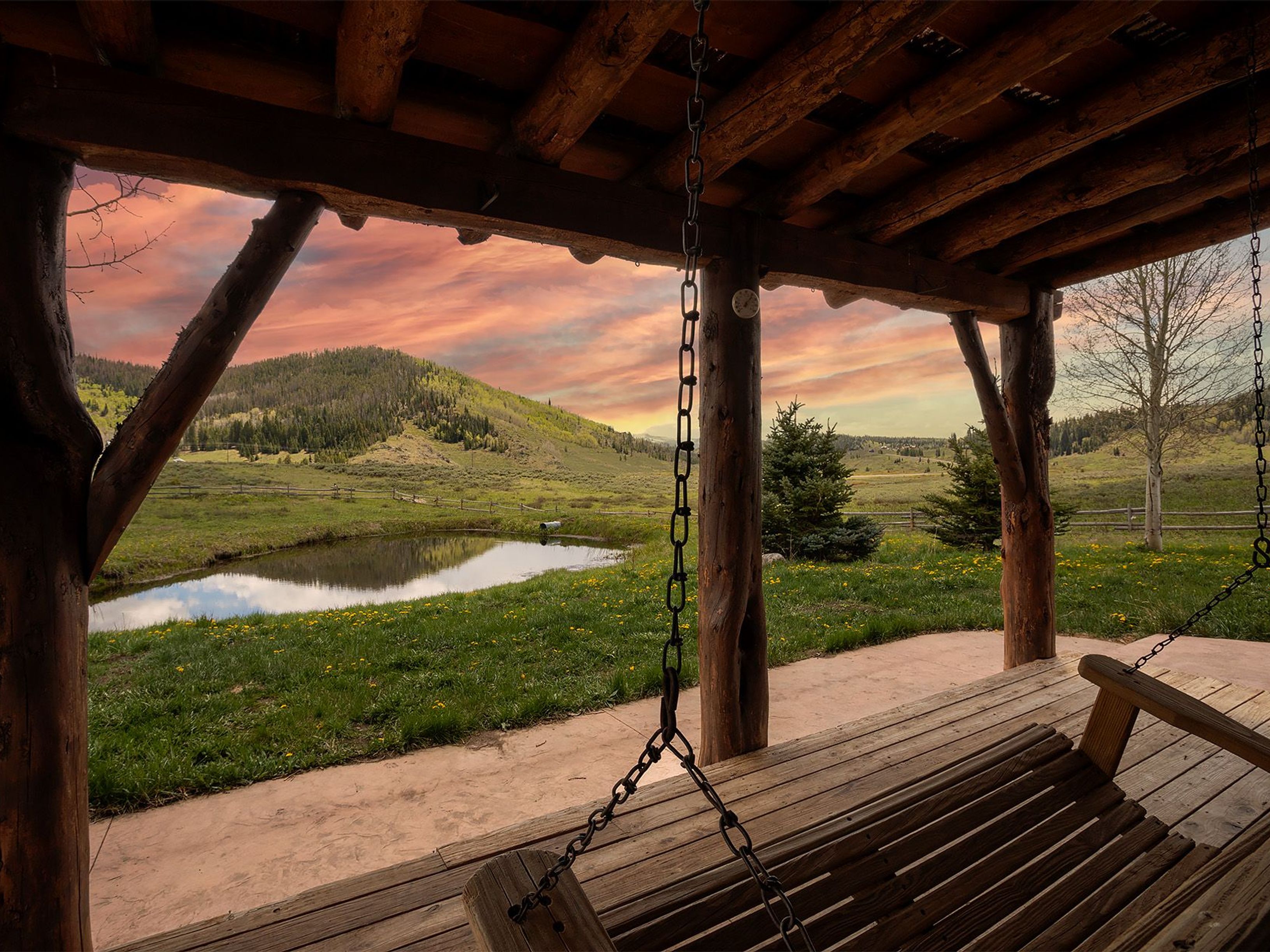 This image showcases a rustic patio or deck with a wooden swing, offering a serene view of a pond and rolling hills under a vibrant sunset sky. The structure is built with sturdy wooden logs, adding to the cabin-like charm. The scene evokes a sense of tranquility and connection with nature, making it an appealing outdoor space for relaxation and enjoying the surrounding landscape.