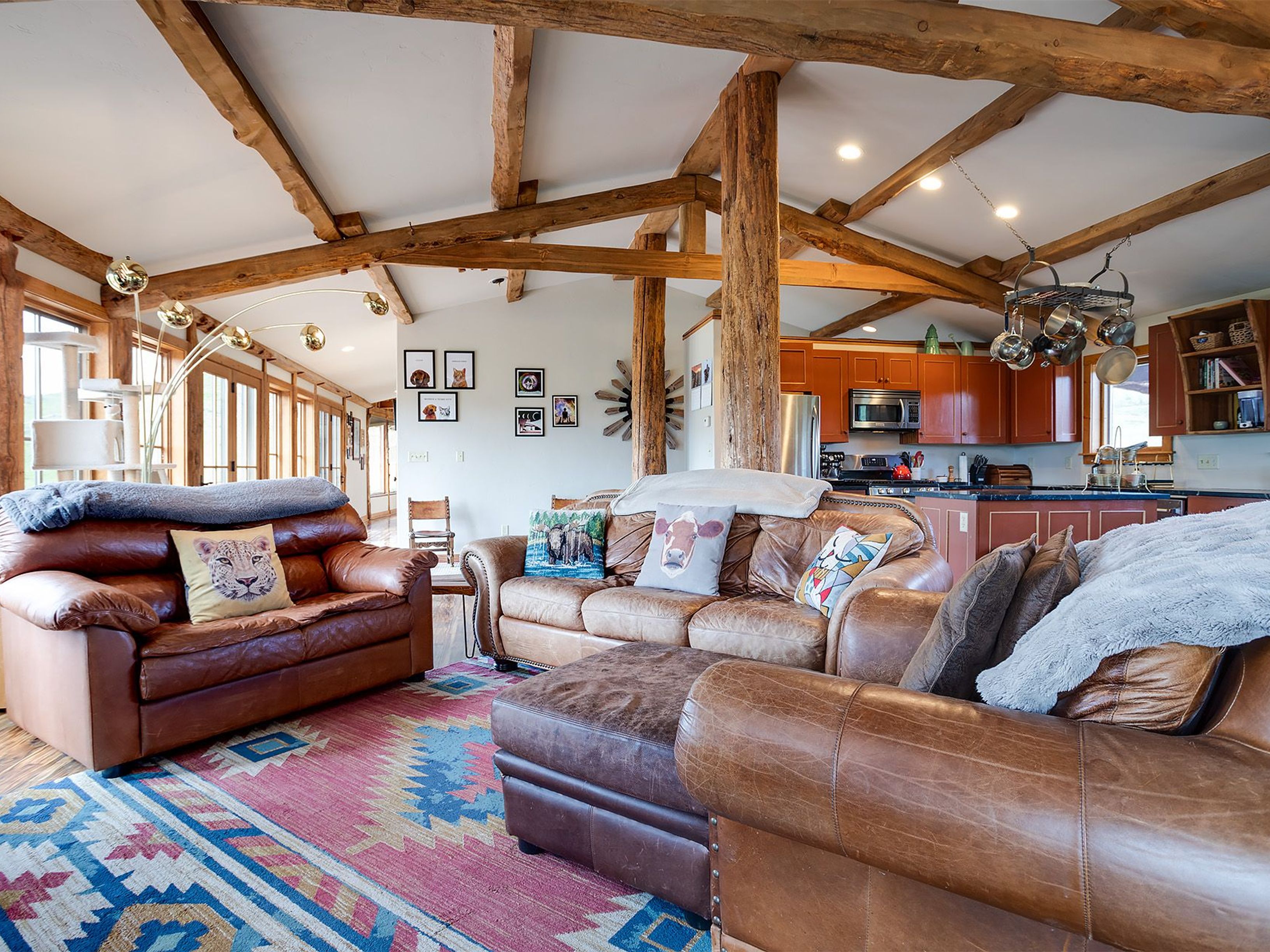This inviting living room features exposed wooden beams and posts, creating a rustic and warm atmosphere. Comfortable leather sofas are arranged around a colorful area rug, with a glimpse into the adjacent kitchen. The space is well-lit and appears spacious, perfect for relaxation and entertaining.