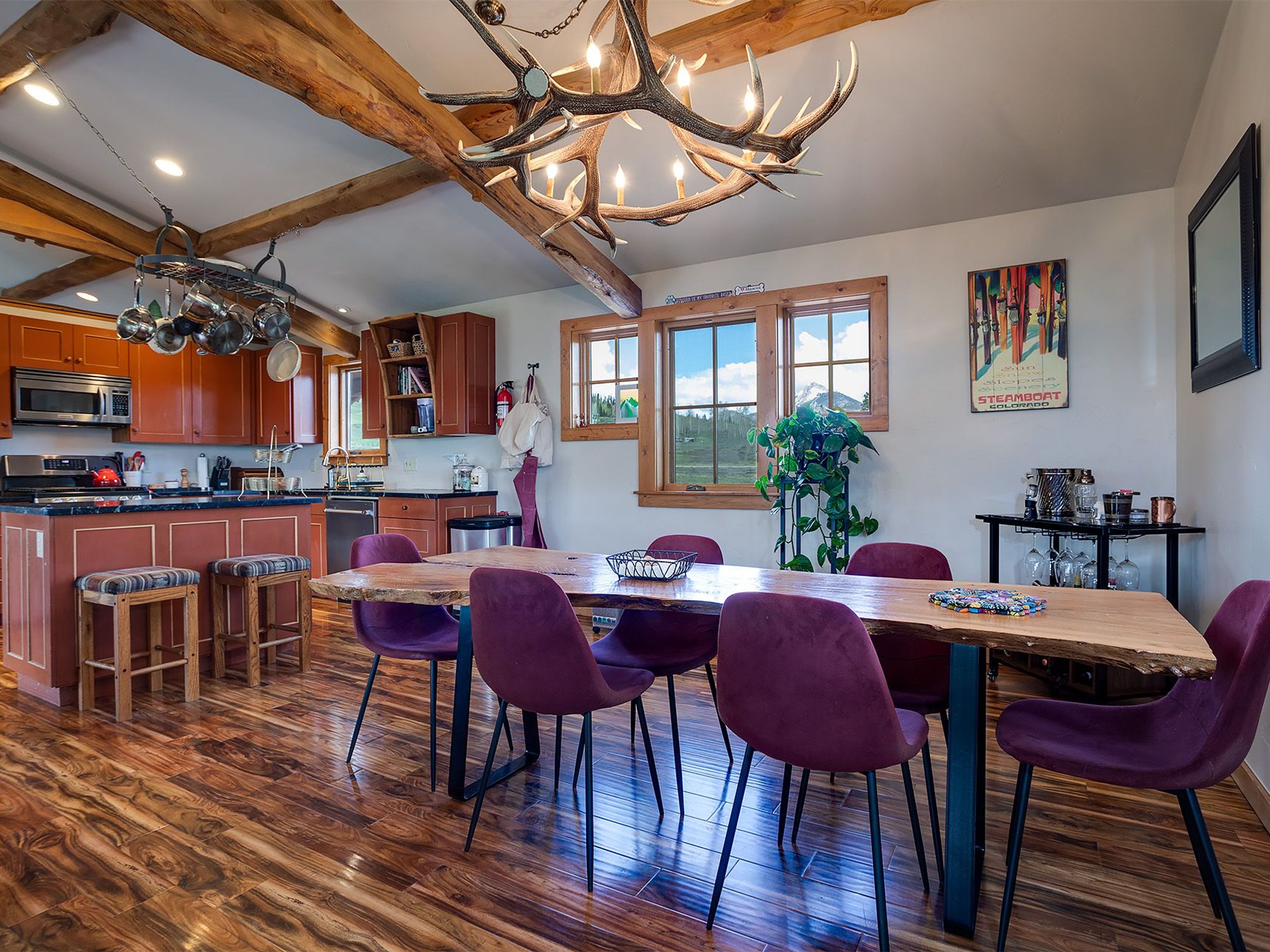 This interior shot showcases a dining area with a rustic-modern aesthetic. A large wooden table with a live edge is surrounded by purple upholstered chairs with black metal legs. An antler chandelier hangs above the table, and the room features exposed wooden beams and hardwood floors, creating a warm and inviting atmosphere.