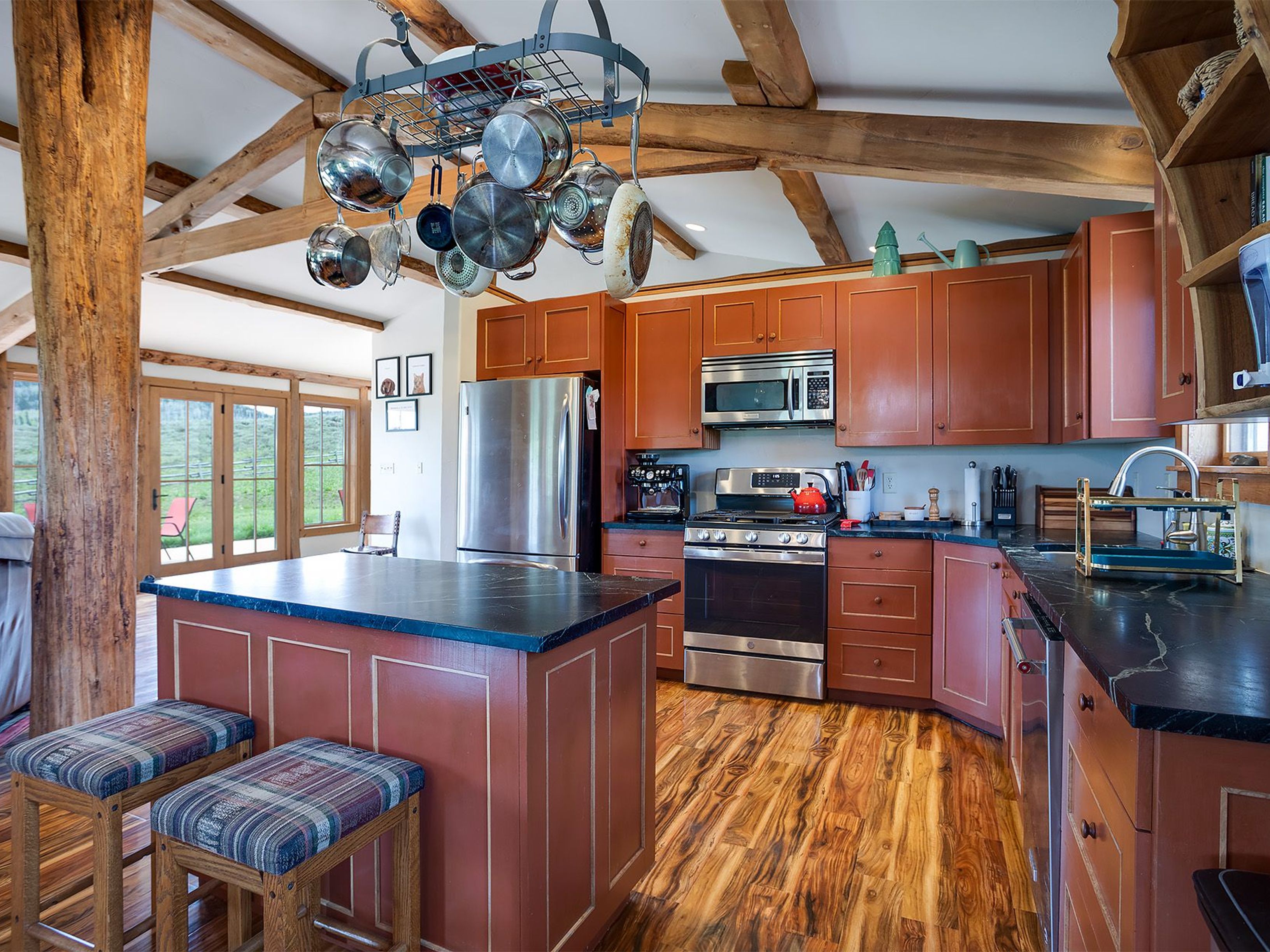 This is a warm and inviting kitchen featuring custom wood cabinetry in a reddish-brown hue with gold trim, complemented by dark countertops and hardwood flooring. A central island with bar seating and a pot rack hanging above the stove add to the rustic charm. The kitchen is well-lit and appears spacious, with a view to the outside through a set of glass doors.