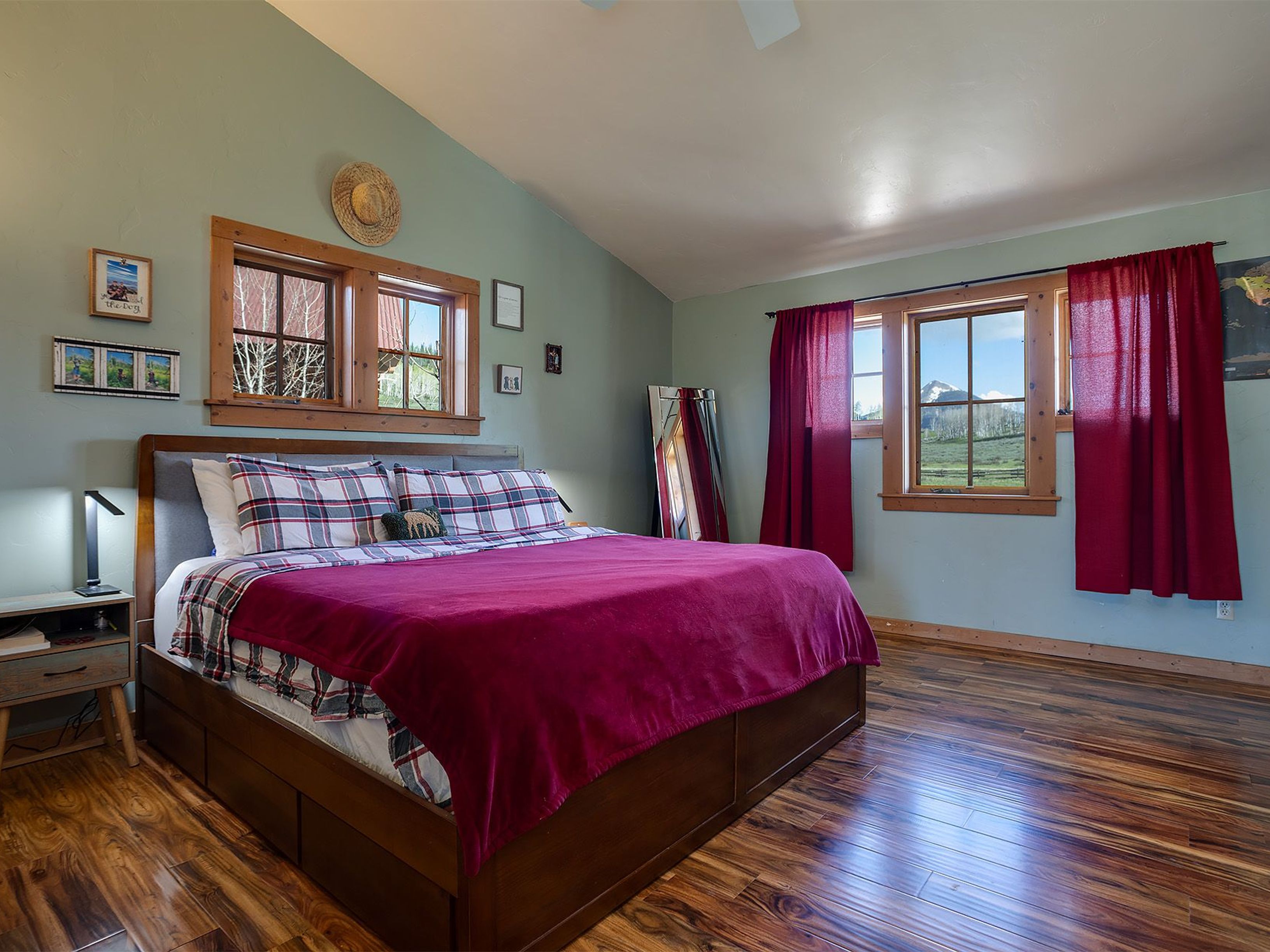 This is a cozy primary bedroom featuring a large bed with a red blanket and plaid pillows. The room has hardwood floors and light green walls, with natural light streaming in through two windows, one with red curtains. The overall impression is warm and inviting, perfect for relaxation.