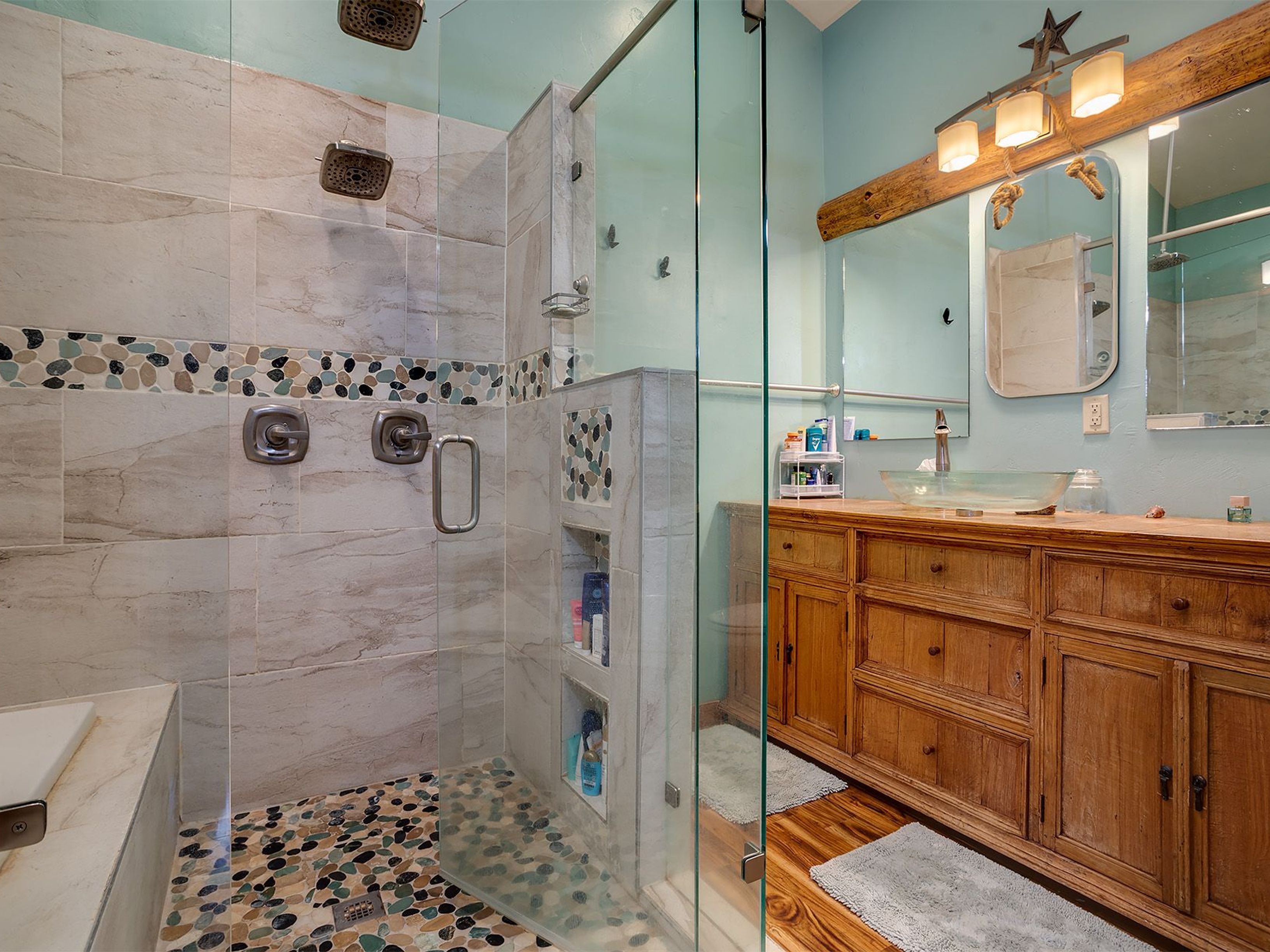 This is a primary bathroom featuring a large walk-in shower with dual shower heads and pebble flooring. The vanity is made of wood with a glass vessel sink. The walls are painted a light blue color, and the overall style is rustic and beachy.
