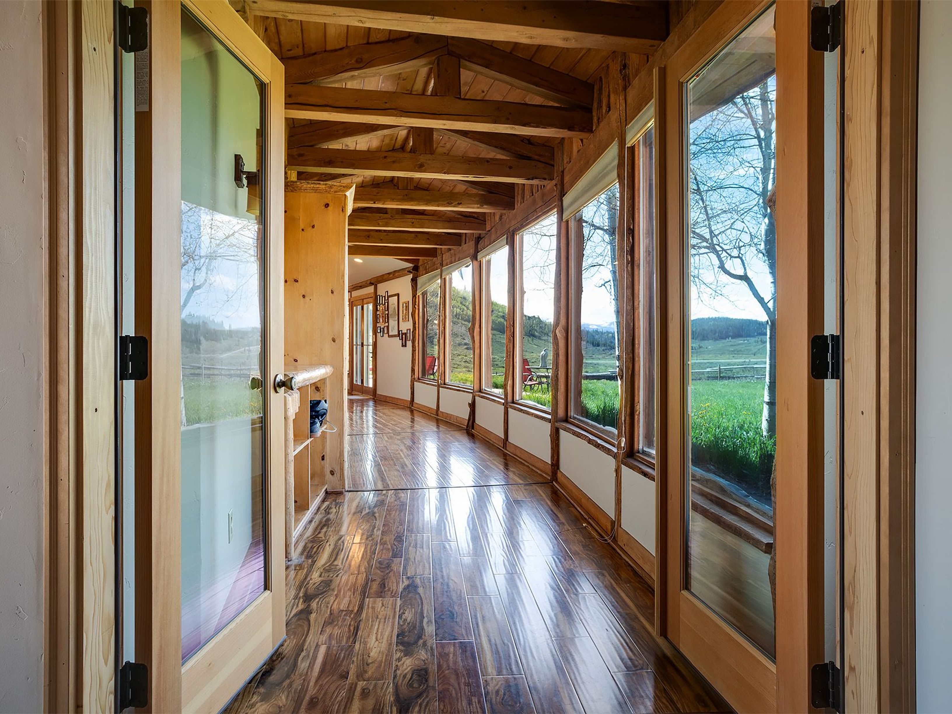 This interior shot showcases a bright hallway with wood-framed glass doors and windows, offering a view of the exterior landscape. The hallway features a wooden ceiling with exposed beams and a glossy hardwood floor, creating a warm and inviting atmosphere. The design emphasizes natural light and a connection to the outdoors, enhancing the home's appeal.