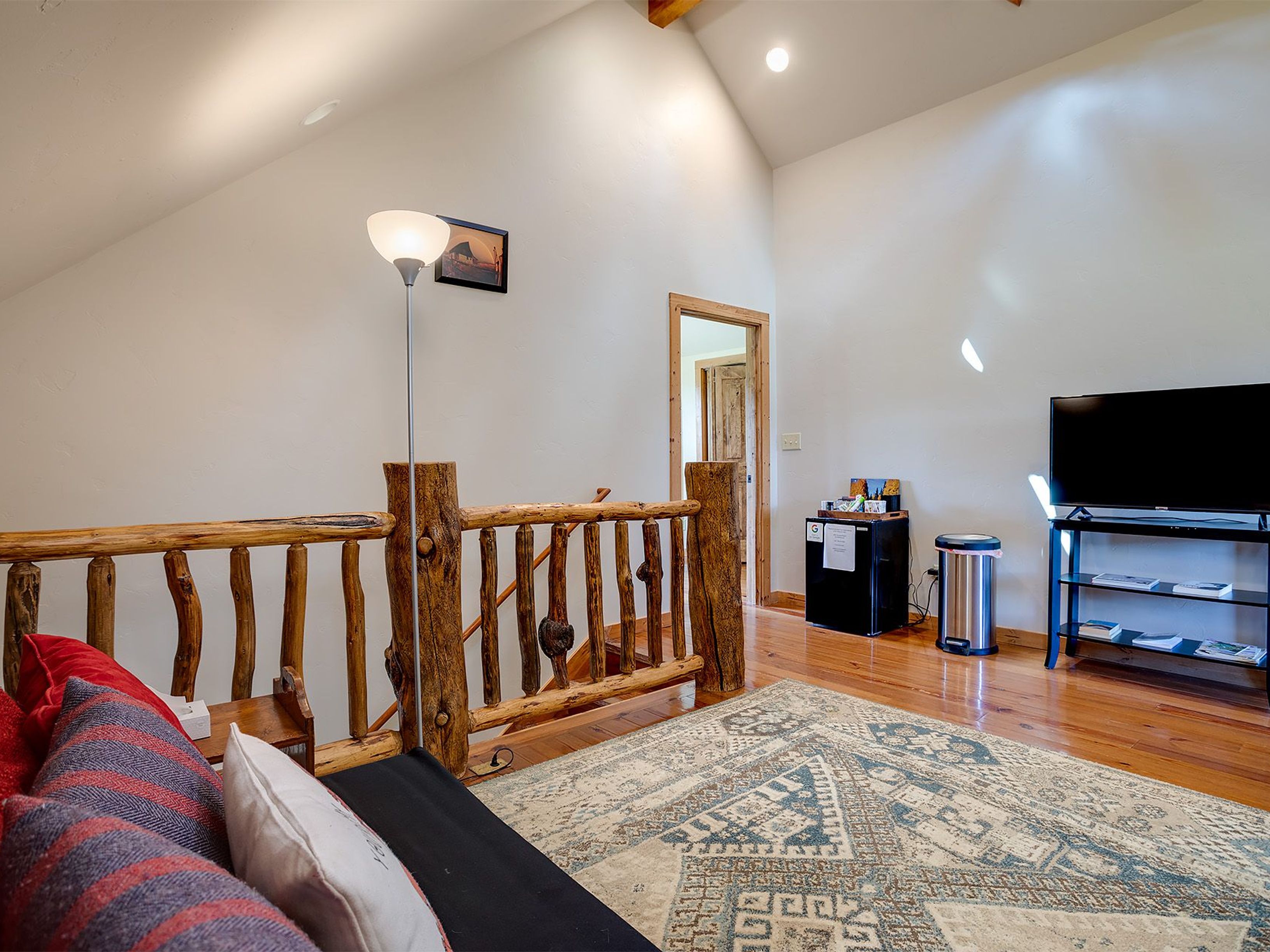 This interior shot showcases a cozy living room area, likely on a second floor or loft, featuring a rustic wooden railing and a comfortable seating area with pillows. A television and mini-fridge are visible, suggesting a relaxed entertainment space. The room is well-lit, with a warm-toned wooden floor and a patterned area rug adding to the inviting atmosphere.