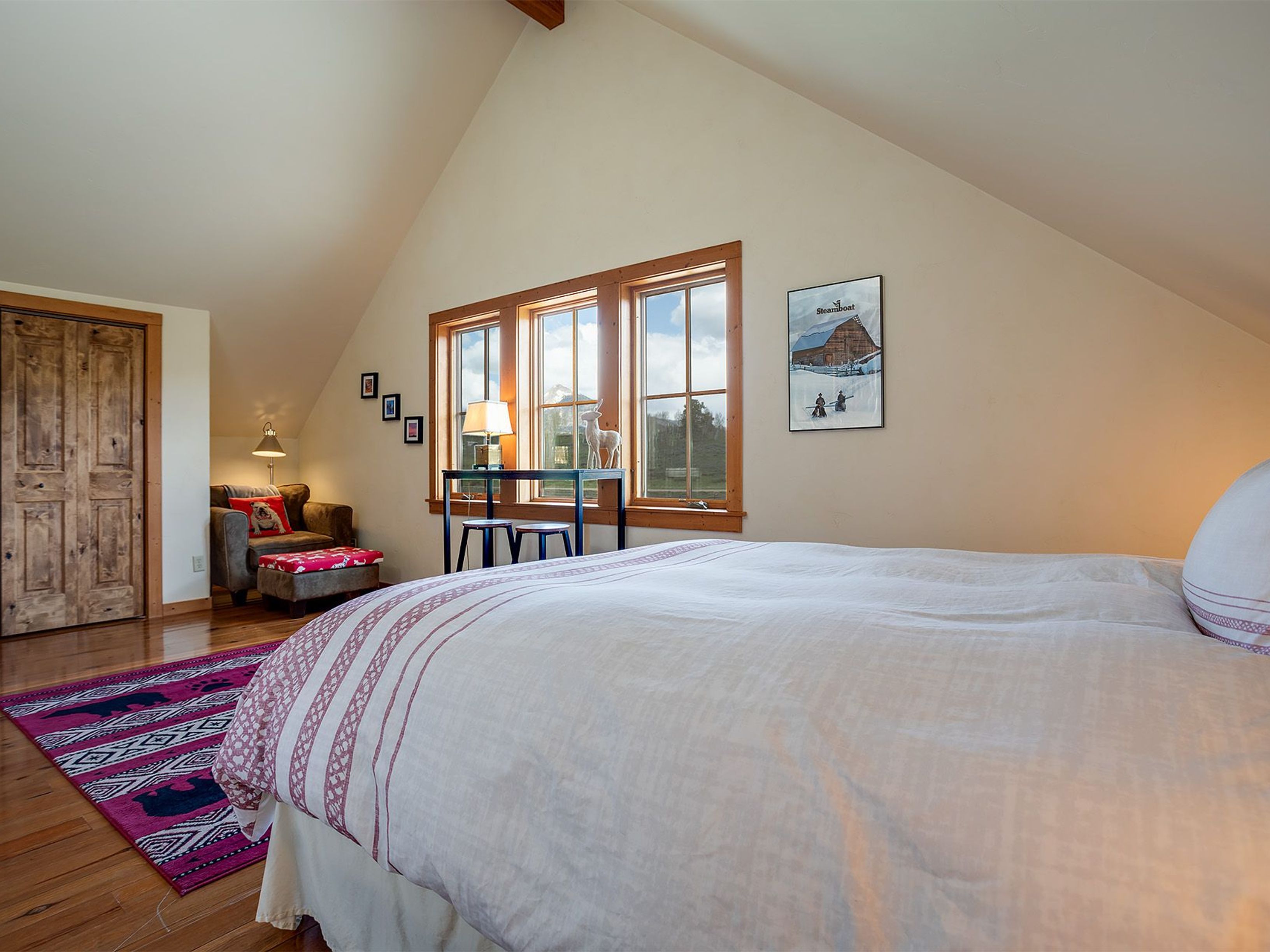 This is a cozy bedroom featuring a large bed with a white and striped duvet, hardwood floors, and a sitting area with an armchair and ottoman. Natural light streams in through the windows, illuminating the room and highlighting the artwork on the walls. The room has a warm and inviting atmosphere, perfect for a guest room or a relaxing retreat.