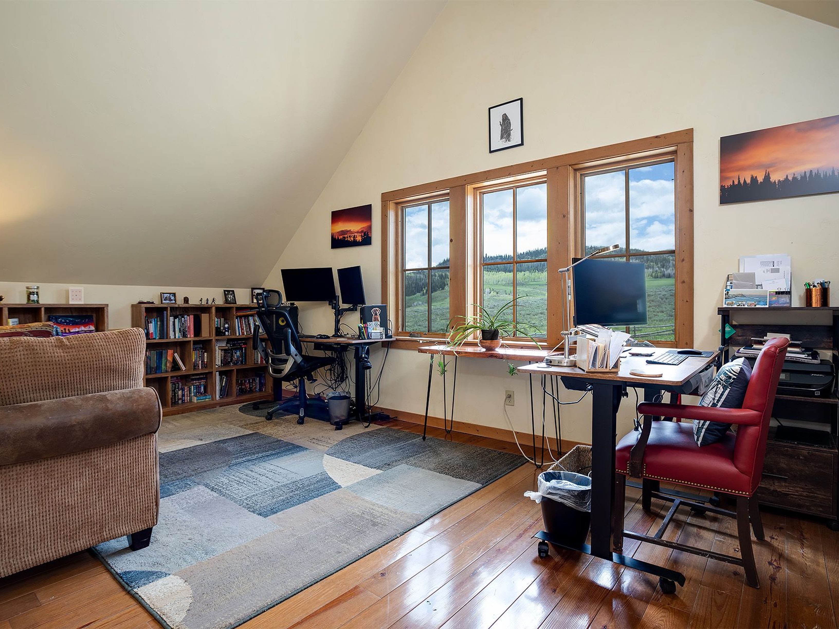 This is an interior shot of a home office featuring hardwood floors and a large area rug. Two desks are positioned in front of a set of three windows offering a view of a green landscape. A bookshelf lines one wall, and a comfortable armchair sits in the corner, creating a cozy and productive workspace.