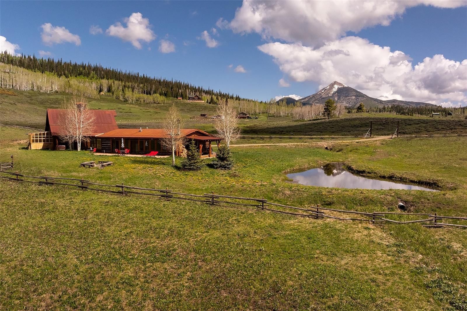This aerial view showcases a charming countryside property featuring a rustic cabin with a distinctive red roof, complemented by a small pond reflecting the sky. The landscape includes rolling green hills, scattered trees, and a backdrop of distant mountains under a partly cloudy sky. A wooden fence adds to the property's character, enhancing its appeal as a serene retreat.