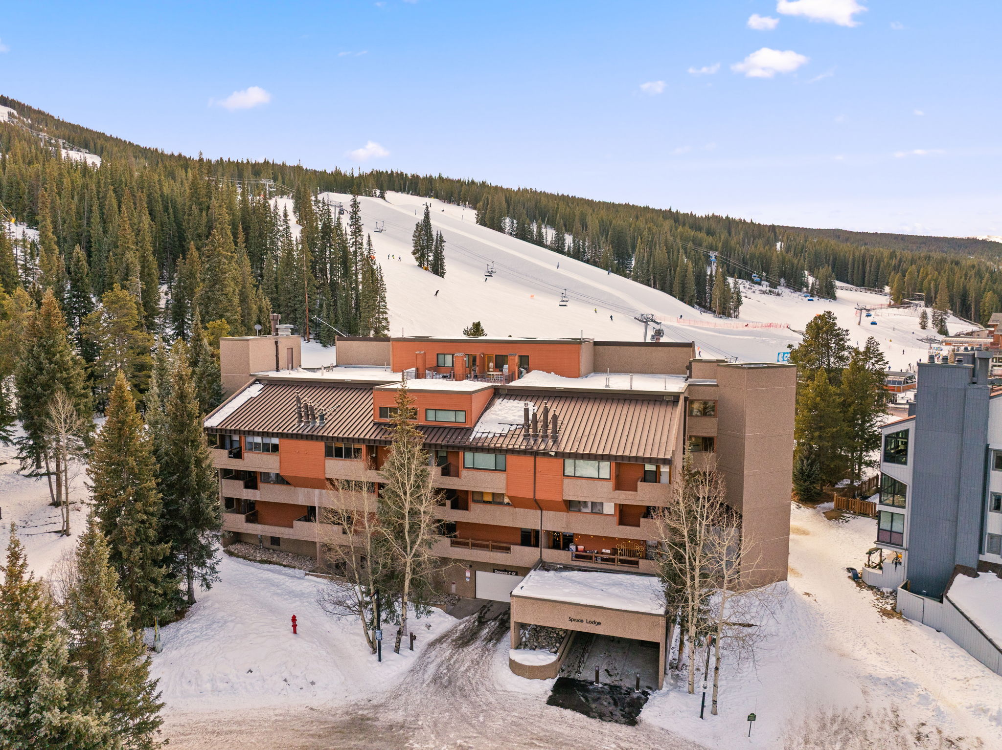 This elevated aerial perspective captures the Spruce Lodge, a multi-story mountain condominium complex nestled at the base of a snow-covered ski slope. The building features a blend of brown and tan architectural elements with a metal roof, surrounded by dense evergreen trees and snowy terrain. The scene conveys a quintessential ski-in, ski-out resort atmosphere, highlighting the property's prime location for winter sports enthusiasts.