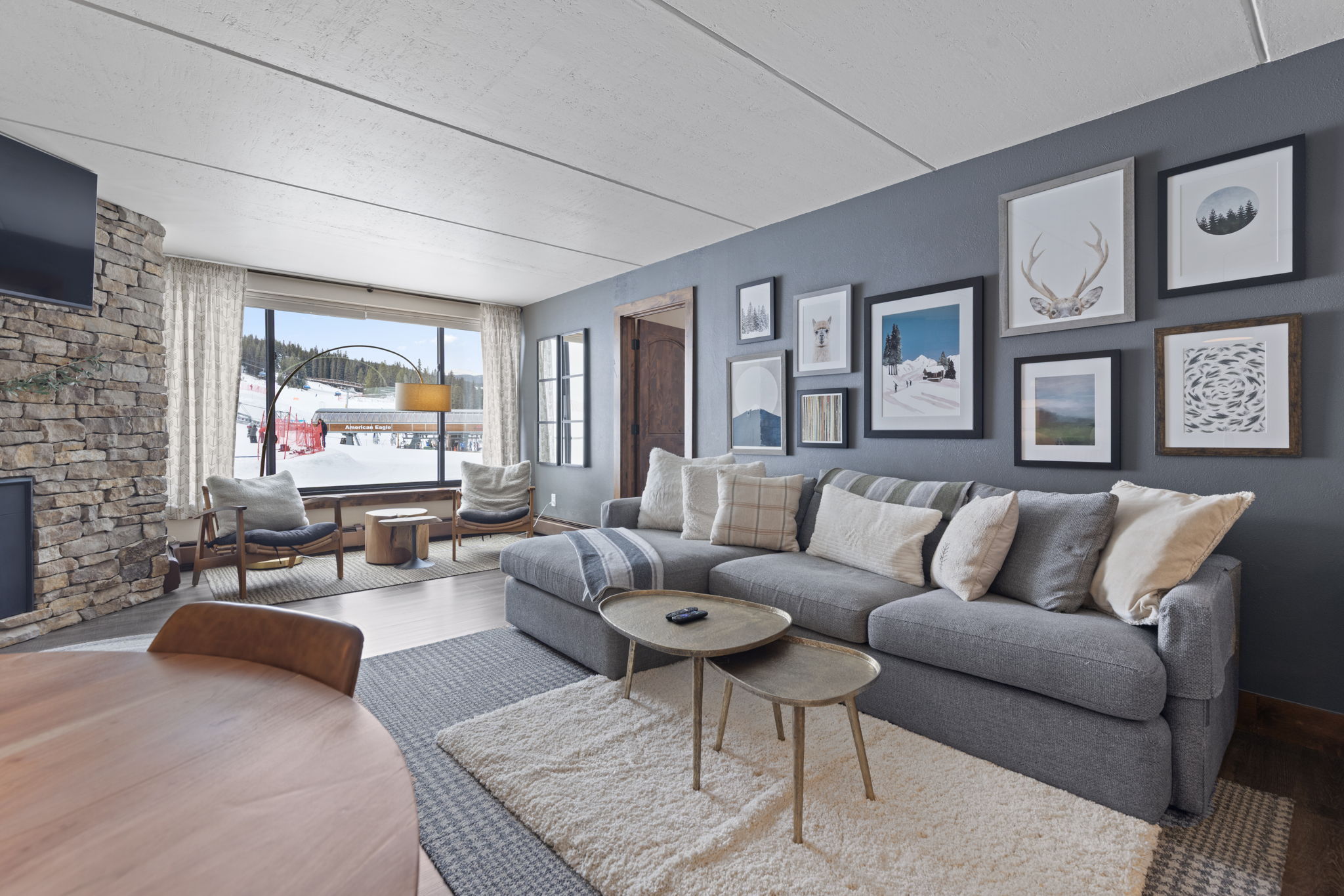 This cozy, mountain-inspired living room features a large grey sectional sofa, a stone fireplace, and a gallery wall of nature-themed artwork. A large window provides a stunning, direct view of a snowy ski slope, creating a perfect après-ski atmosphere. The space is anchored by a plush area rug and layered coffee tables, blending rustic charm with modern comfort.