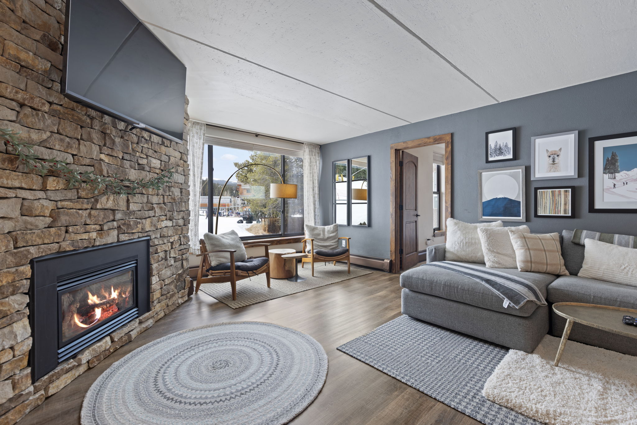 This cozy, mountain-inspired living room features a striking floor-to-ceiling stone fireplace with an integrated television and a warm, inviting fire. The space is furnished with a plush grey sectional sofa, two mid-century modern armchairs, and layered area rugs, all set against a backdrop of soft grey walls and a large window overlooking a snowy landscape. The room exudes a comfortable, rustic-chic atmosphere perfect for relaxing after a day on the slopes.