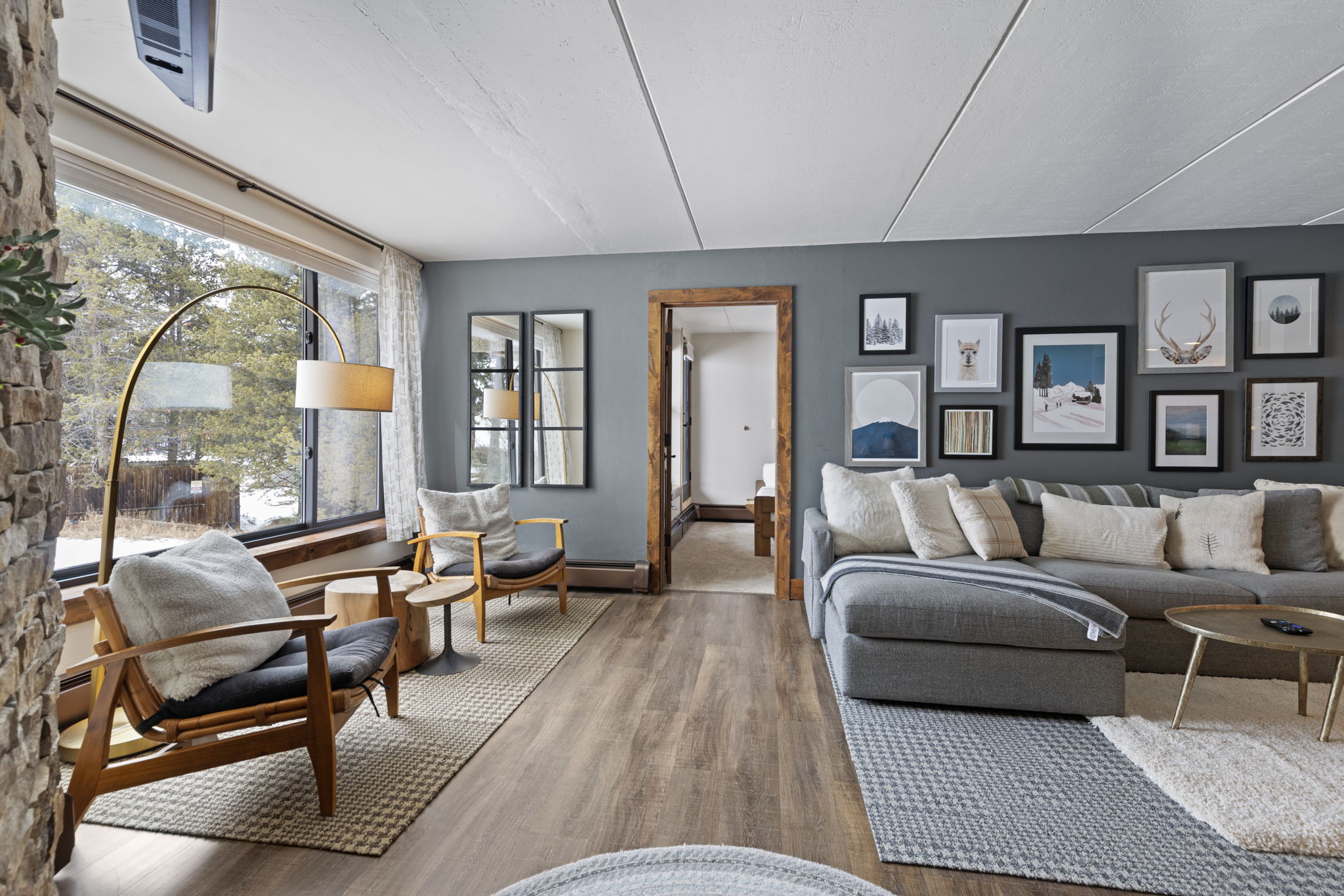 This inviting living room features a cozy, modern aesthetic with a large grey sectional sofa, a gallery wall of framed art, and two mid-century modern armchairs positioned by a large window. The space is anchored by textured area rugs over wood-look flooring, with a stone accent wall and a warm, natural light filtering in from the outdoors. The perspective is a wide-angle shot that captures the comfortable, open-concept flow of the living area.