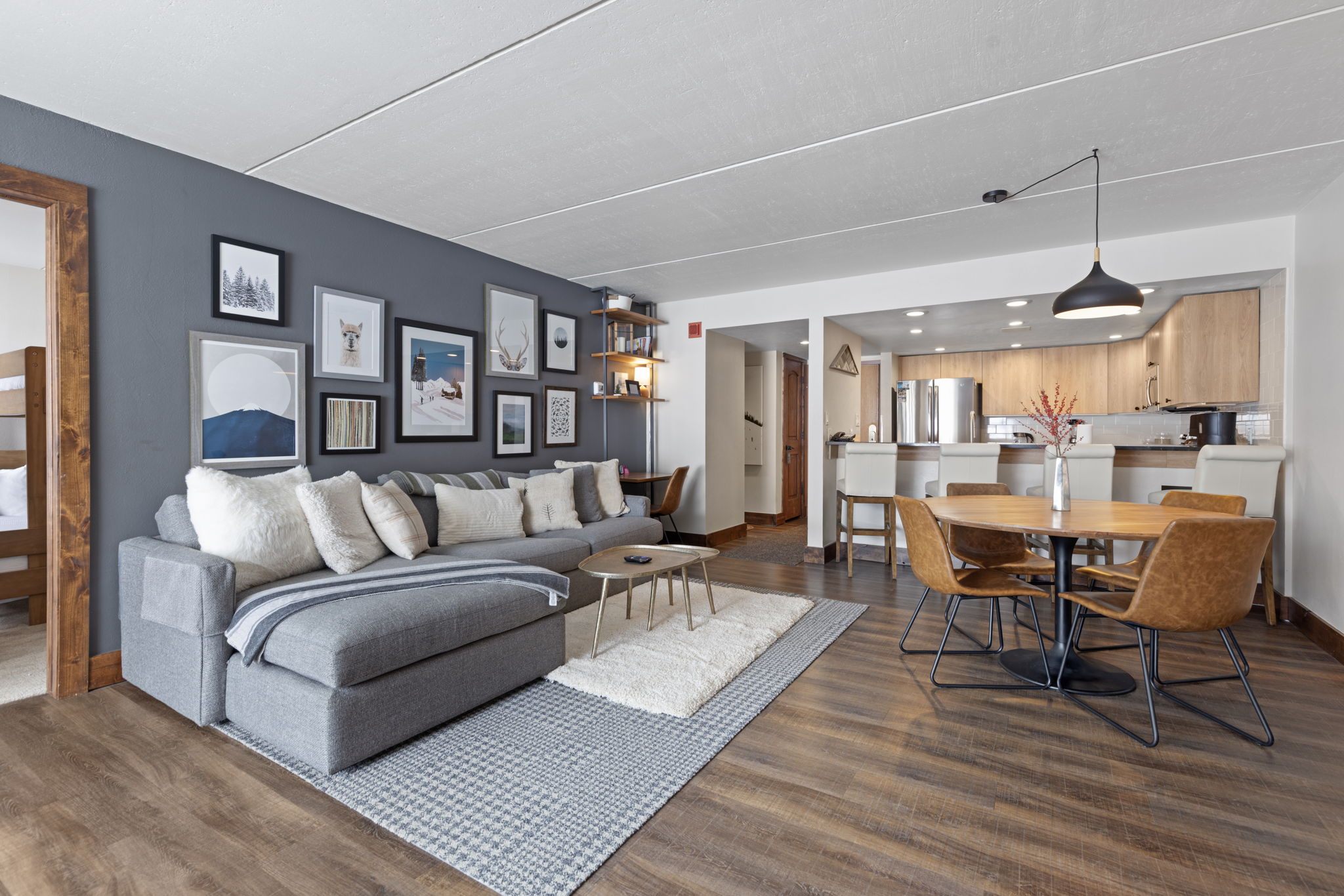 This open-concept living and dining area features a cozy, modern aesthetic with a large grey sectional sofa, a gallery wall of framed art, and warm wood-look flooring. The space flows seamlessly into a dining area with a round table and leather chairs, adjacent to a kitchen with light wood cabinetry and a breakfast bar. The perspective is wide and inviting, showcasing a well-lit, functional layout perfect for comfortable living.